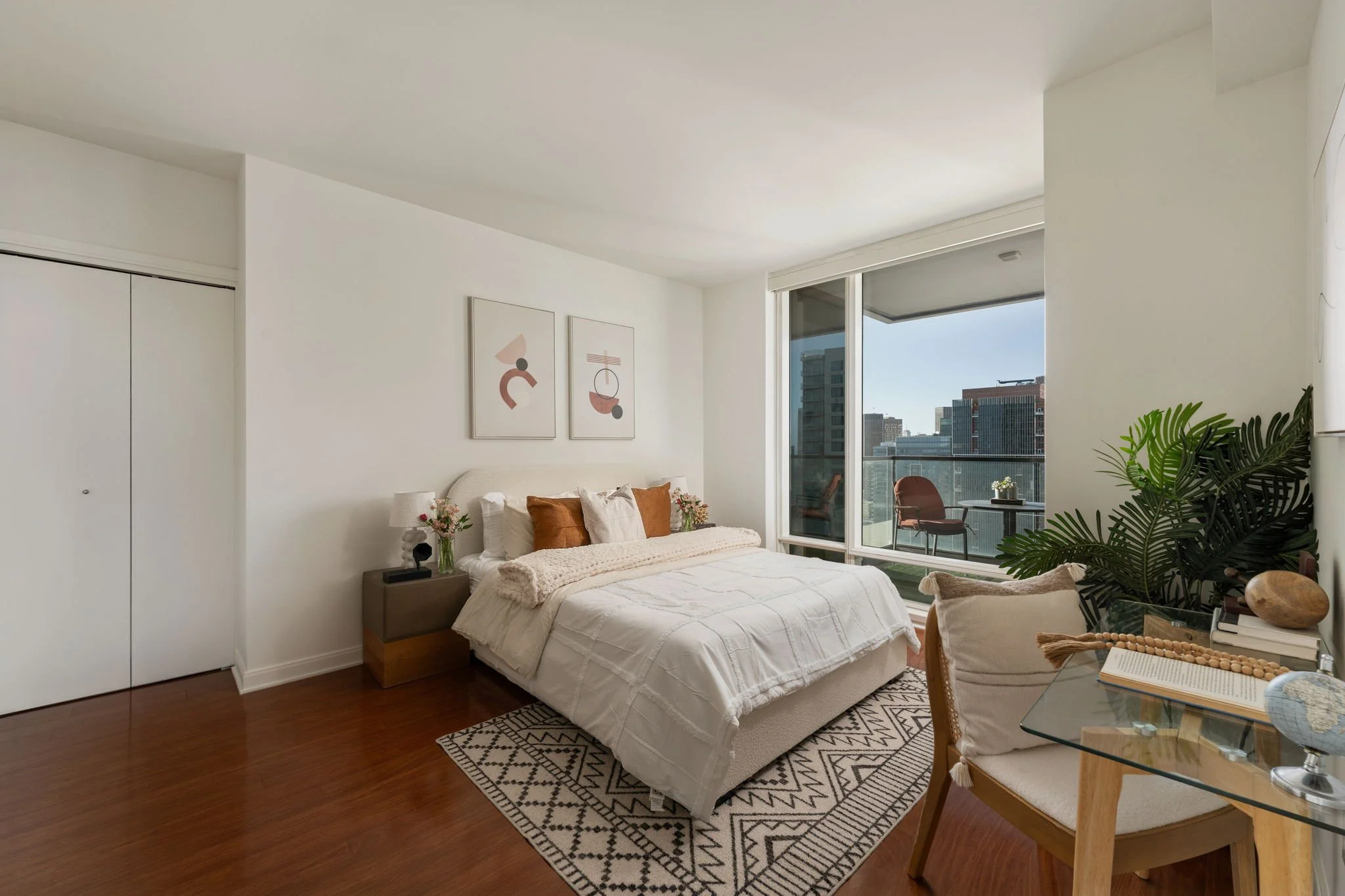 Modern bedroom with a large bed, nightstand with a lamp and flowers, artwork on the wall, large window with a balcony view, and a dining table with chairs, plant, and decorative items.