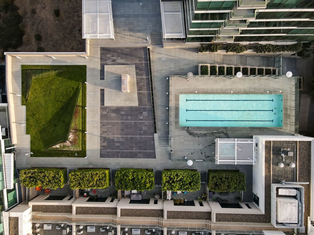 An aerial view of a modern residential building's rooftop featuring a swimming pool, a grassy area, a sitting area, and landscaping.