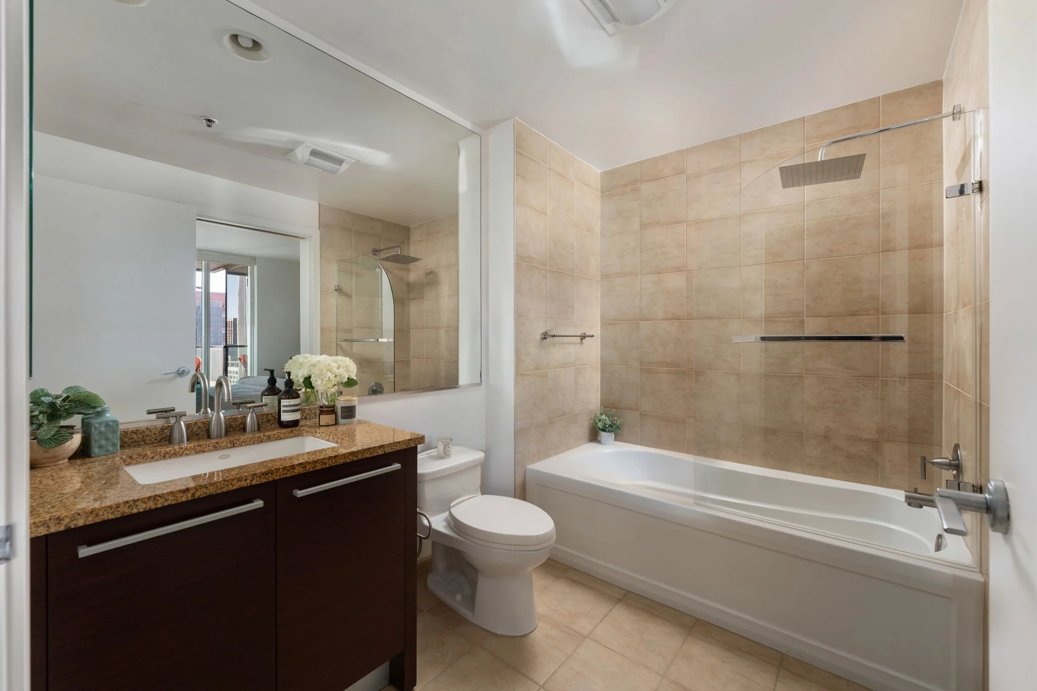 Bathroom with a vanity with a granite countertop, a sink, soap dispensers, a potted plant, a mirror, a toilet, and a bathtub with a glass shower cover, beige tiled walls, and a ceiling with exhaust fan and lighting.