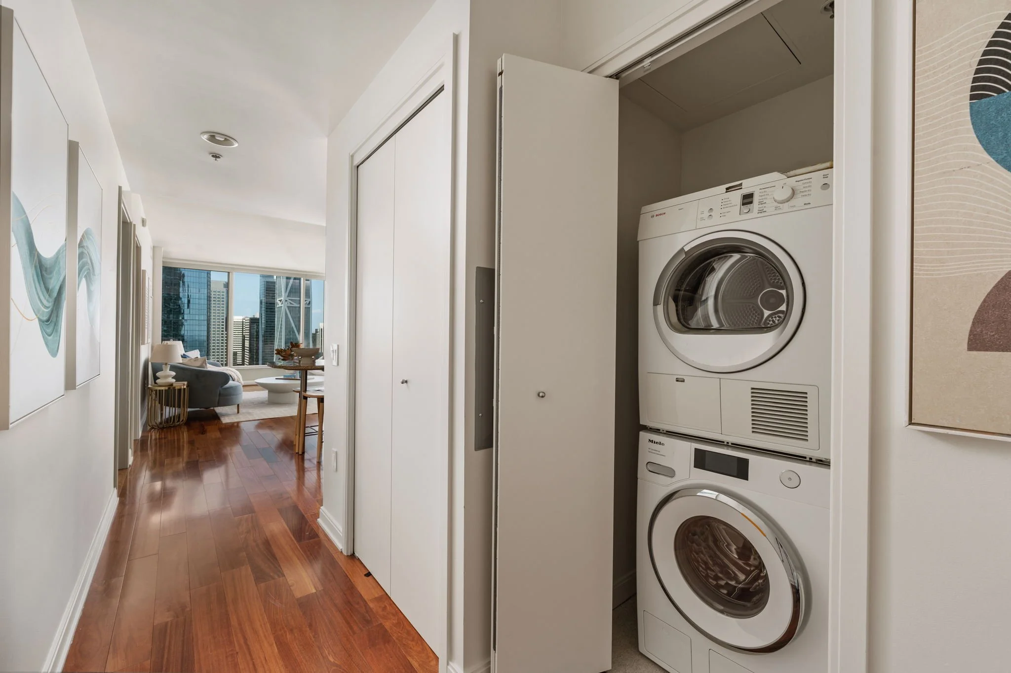 View of a laundry area with stacked washer and dryer inside a closet, with a hallway leading to a living room with large windows and modern cityscape view.
