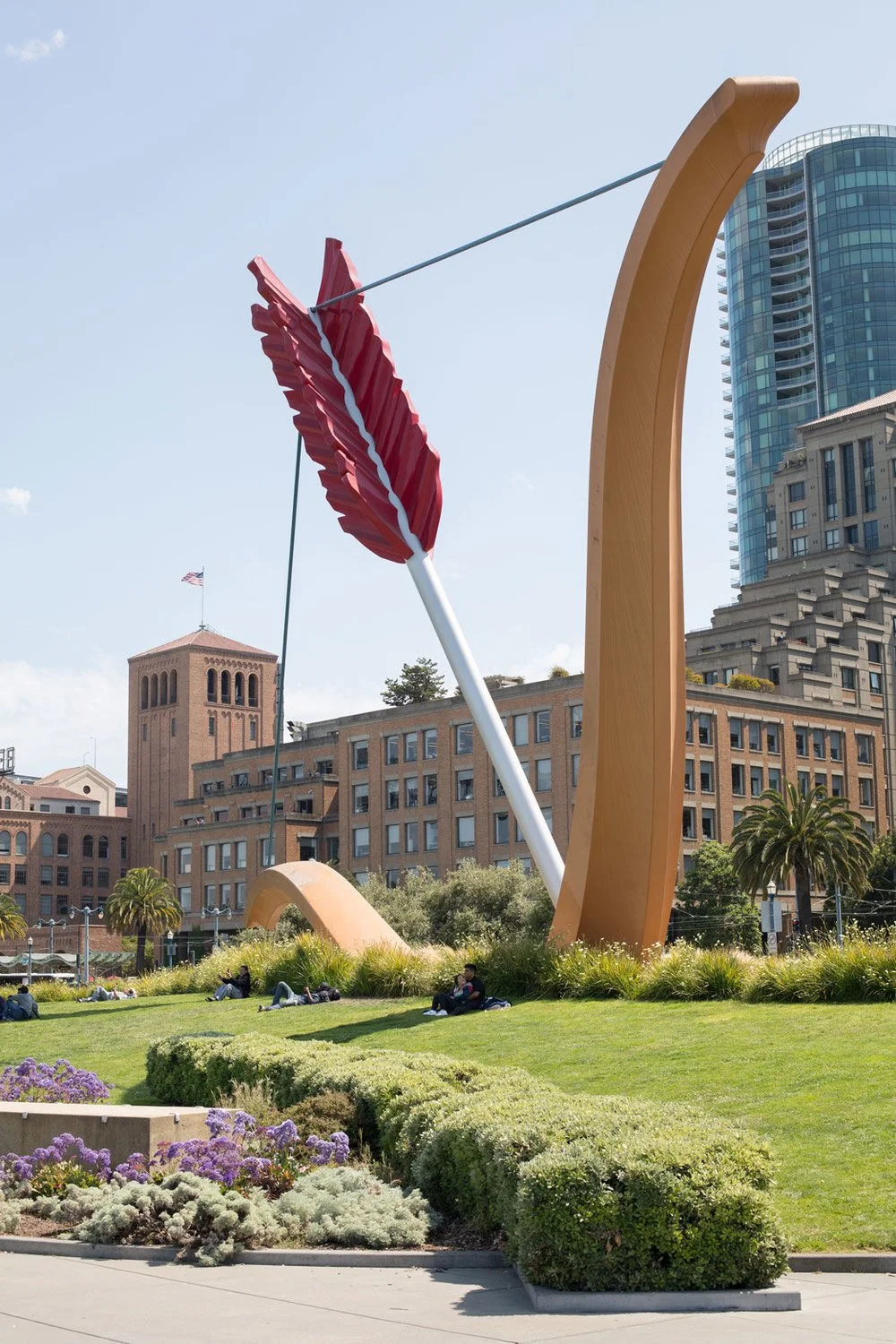 A large sculpture of a bow and arrow in a park with buildings in the background. The arrow has a red feather, and the bow is beige, surrounded by greenery and purple flowers.