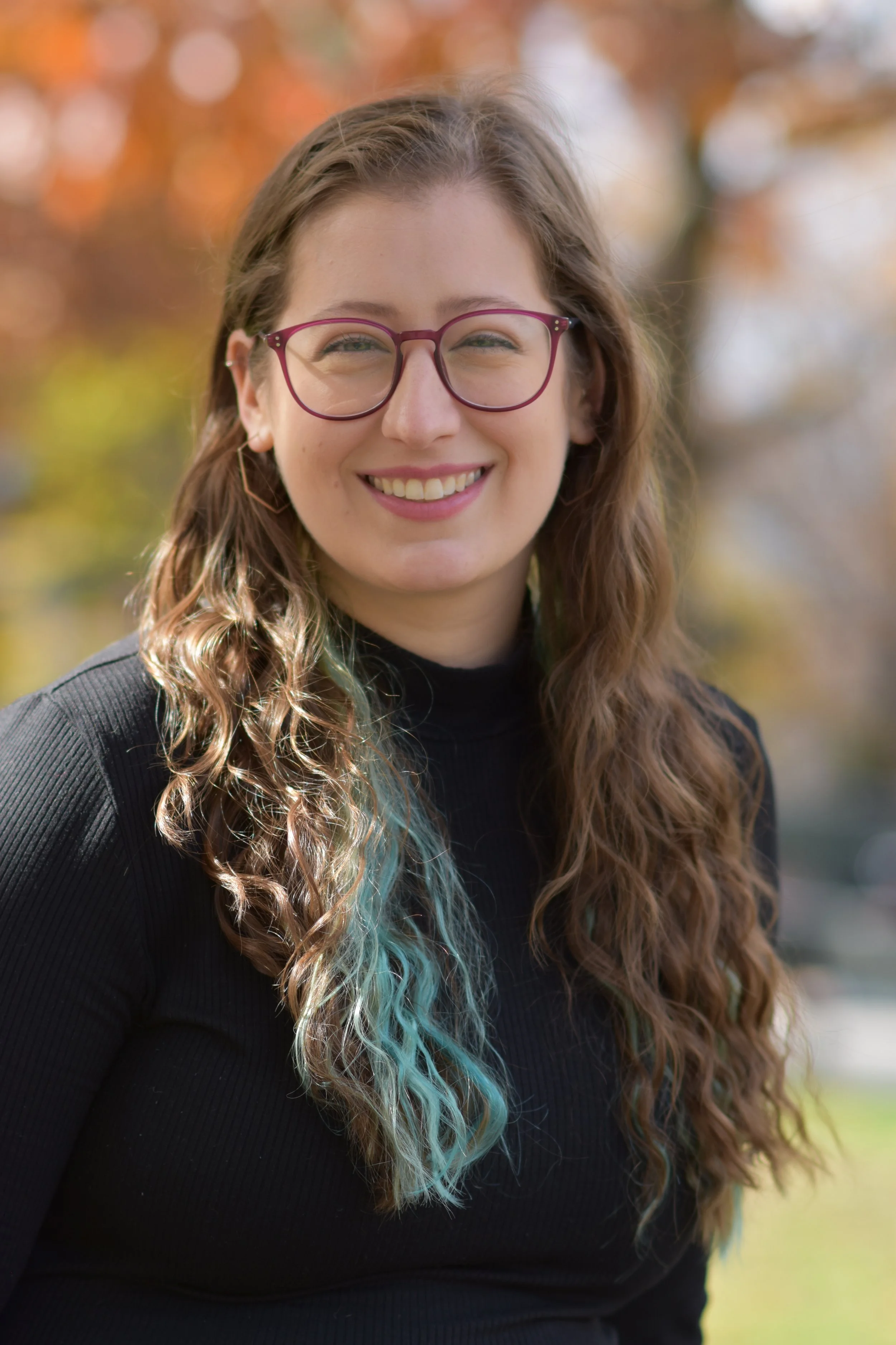 Jess is a white woman with long wavy brown hair with a blue streak, glasses, and a welcoming smile.