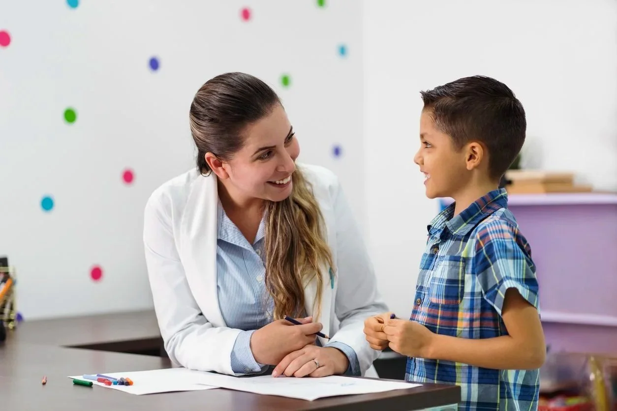 Speech Therapy talking with a young boy at a table with papers and colored markers, working on improving his communication skills during speech therapy for kids in Honolulu, HI