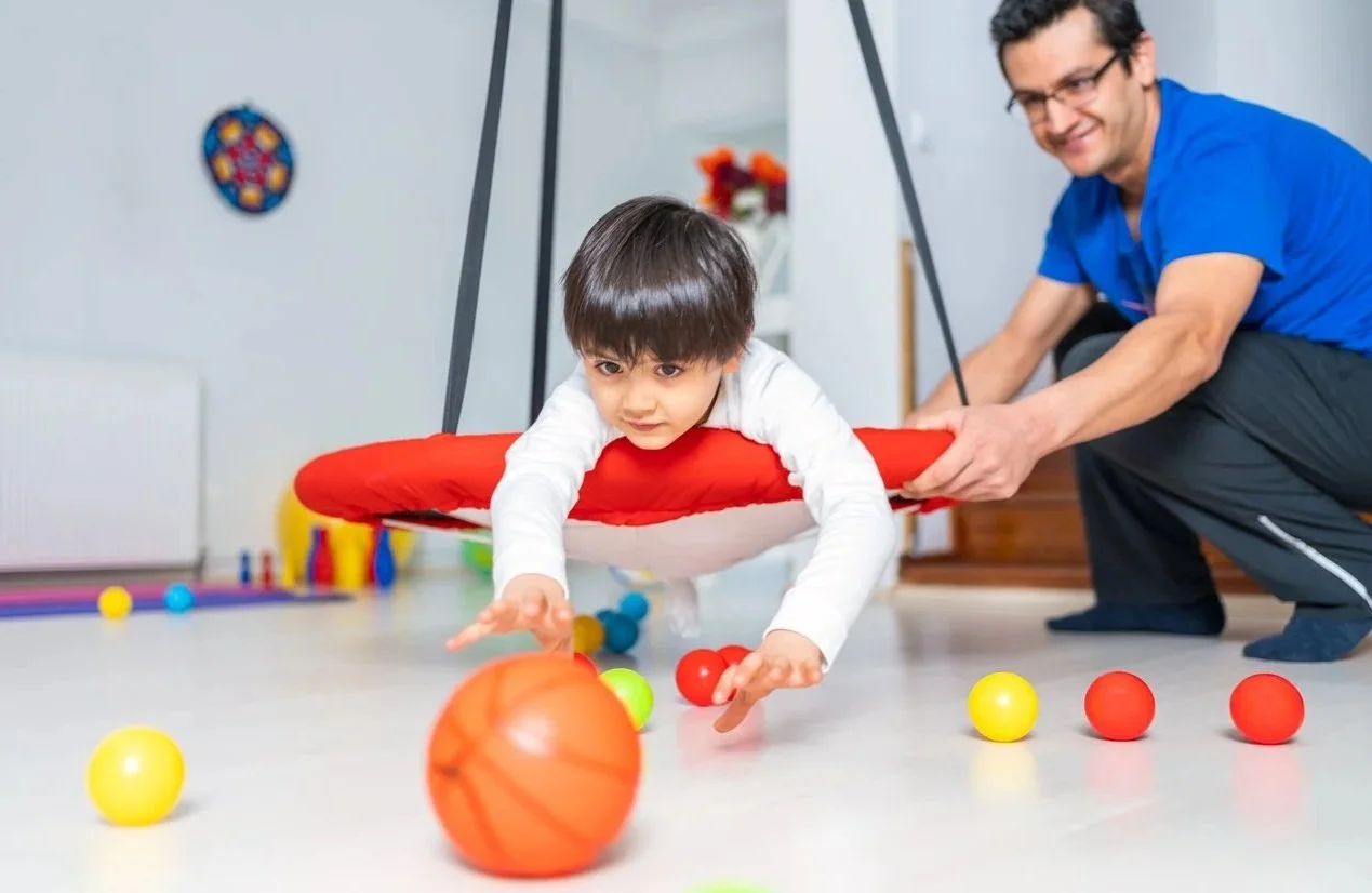 Child playing in a sensory swing while a physical therapist assists, surrounded by colorful balls indoors. The child is working on sensory regulation during occupational therapy in Honolulu, HI