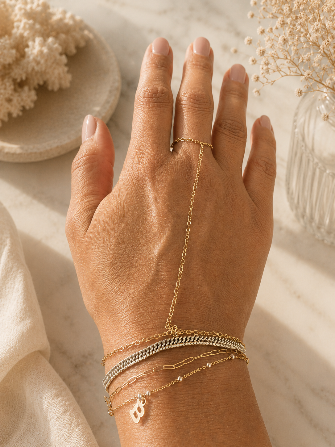 A person's hand with multiple gold bracelets and a gold ring featuring a dangling chain, against a neutral background with decorative dried flowers.