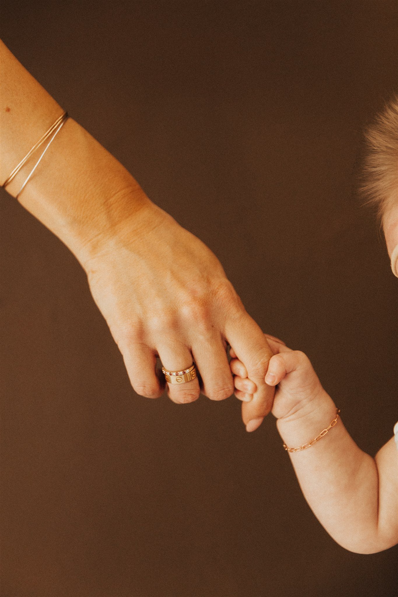 A hand of an adult with a wedding band holding the hand of a child with a bracelet, both with rings on their fingers, against a dark background.