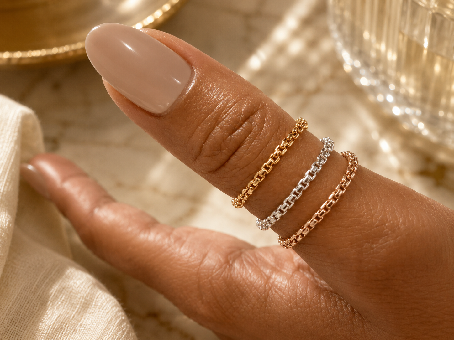 A close-up of a person's hand displaying three delicate chain rings on the fingers, in gold, silver, and rose gold tones, with a blurred background of glassware and warm lighting.