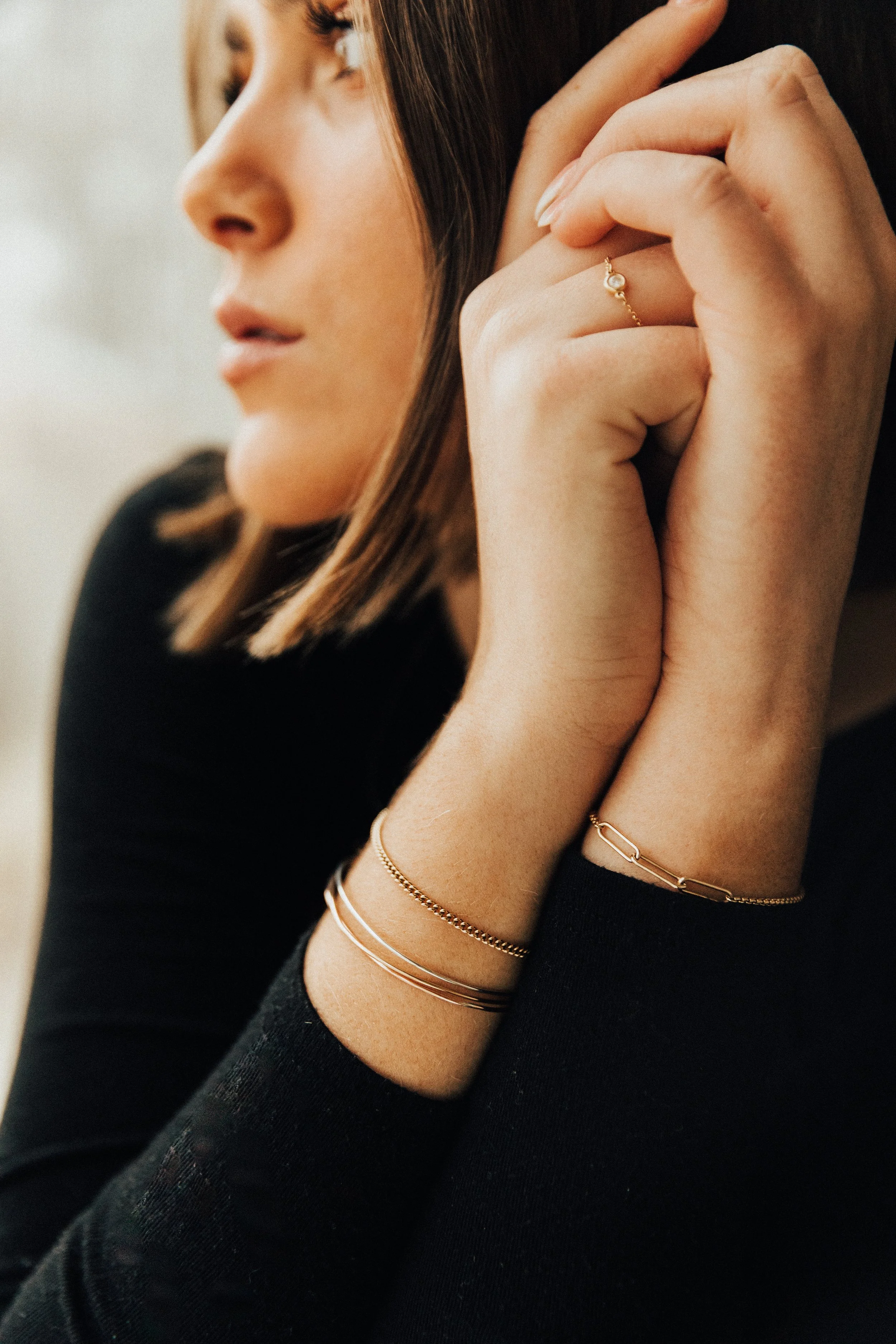 Close-up of a woman with fair skin and brown hair, wearing black clothing, gold jewelry, and a ring on her finger, touching her face and looking to the side.