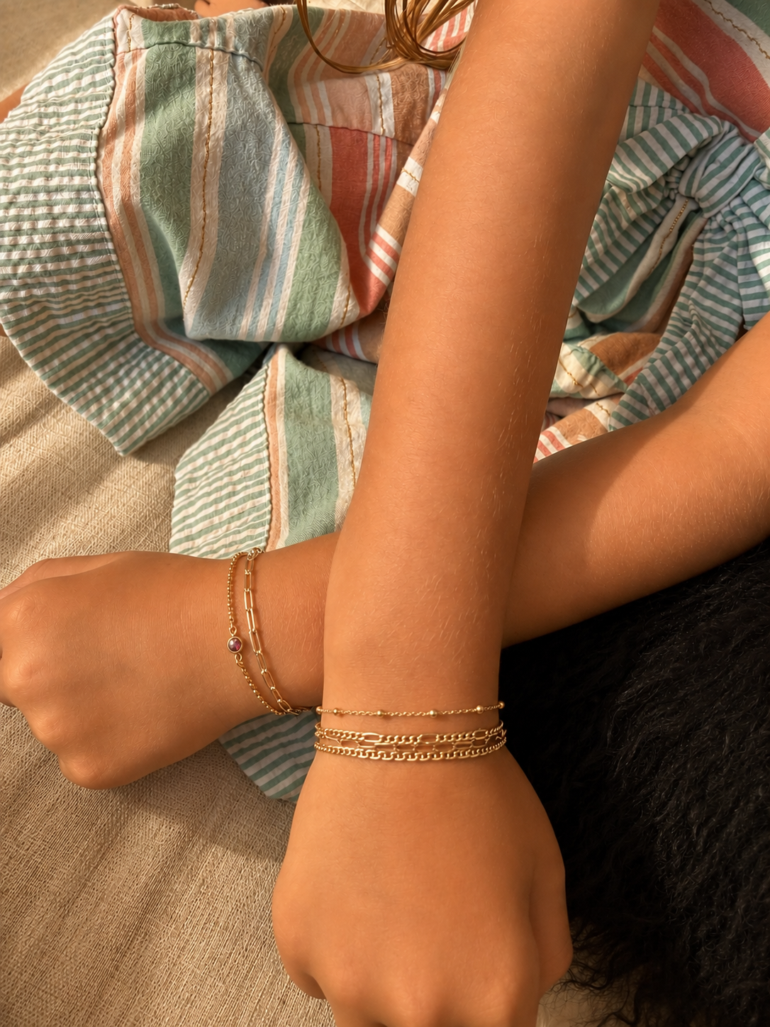 Close-up of a person's arm resting on their knee, wearing multiple gold bracelets. They are lying on a bed with pastel-colored, striped, and textured bed sheets.