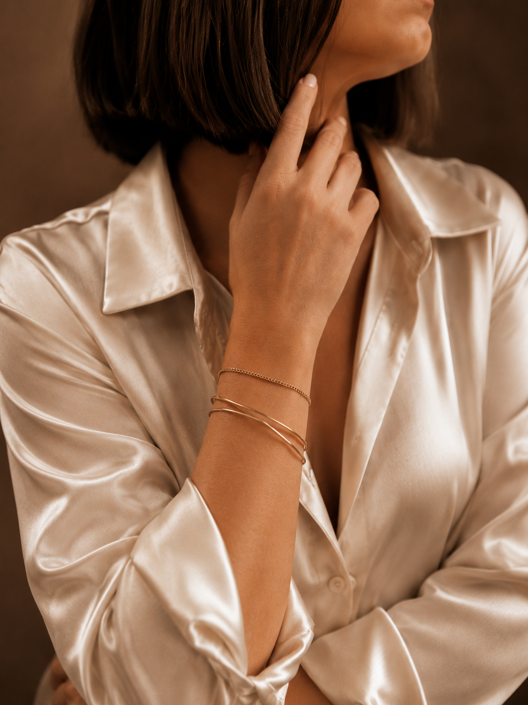 Close-up of a woman with shoulder-length brown hair wearing a shiny silk cream-colored shirt, with her hand touching her neck, wearing a delicate gold bracelet and chain.