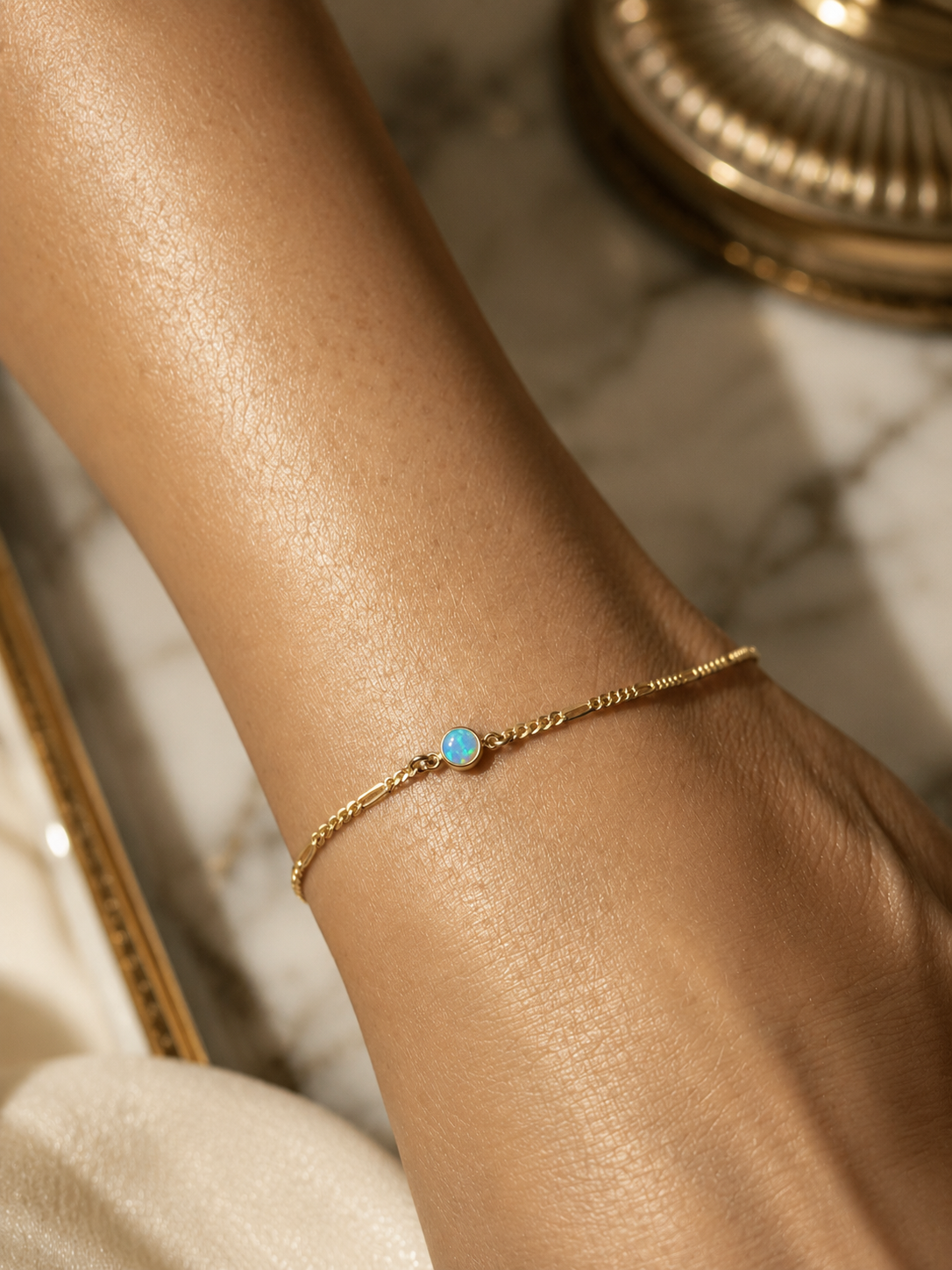 A gold chain bracelet with an opal stone on a person's wrist, with a marble surface and a decorative metal object in the background.