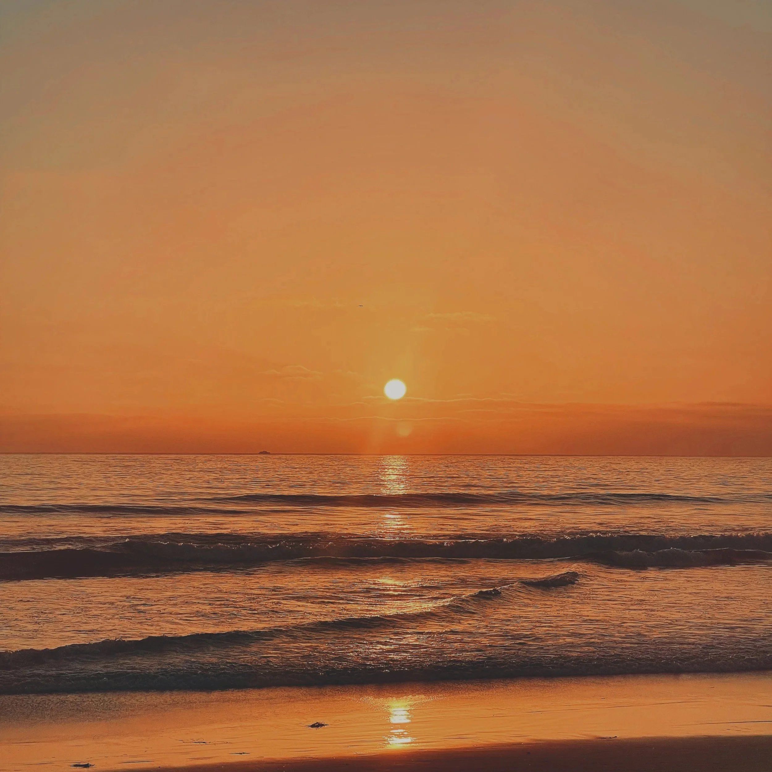 Sunset over the ocean with the sun near the horizon, casting a golden glow on the water and sky, with gentle waves at the shoreline.