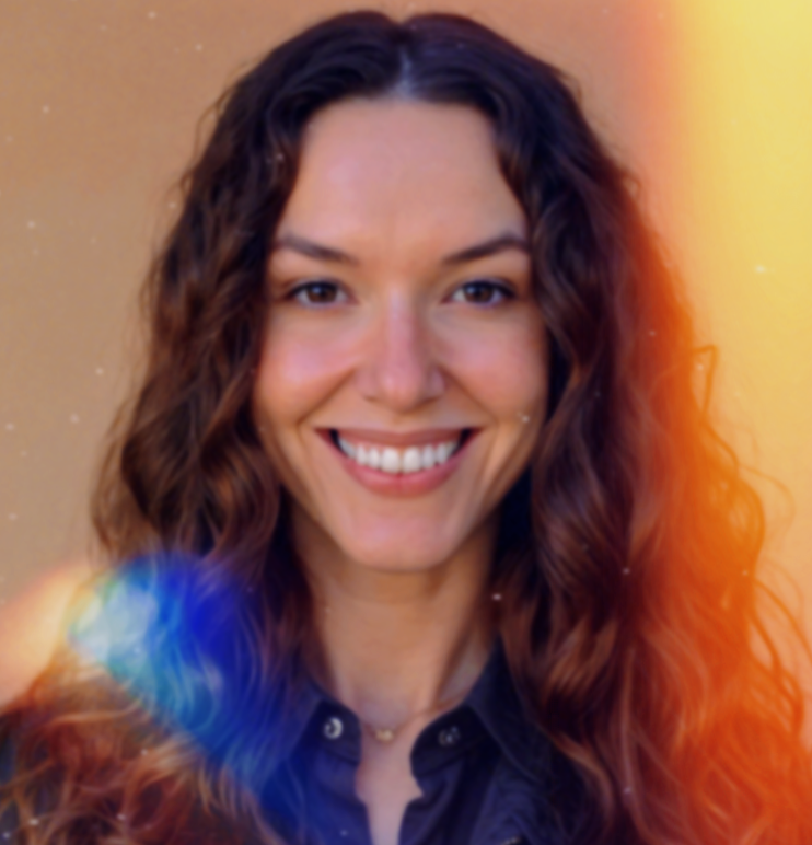 A woman with long, curly brown hair smiling at the camera, wearing a dark shirt with a gold necklace, with a colorful light flare and a gradient yellow-orange background.