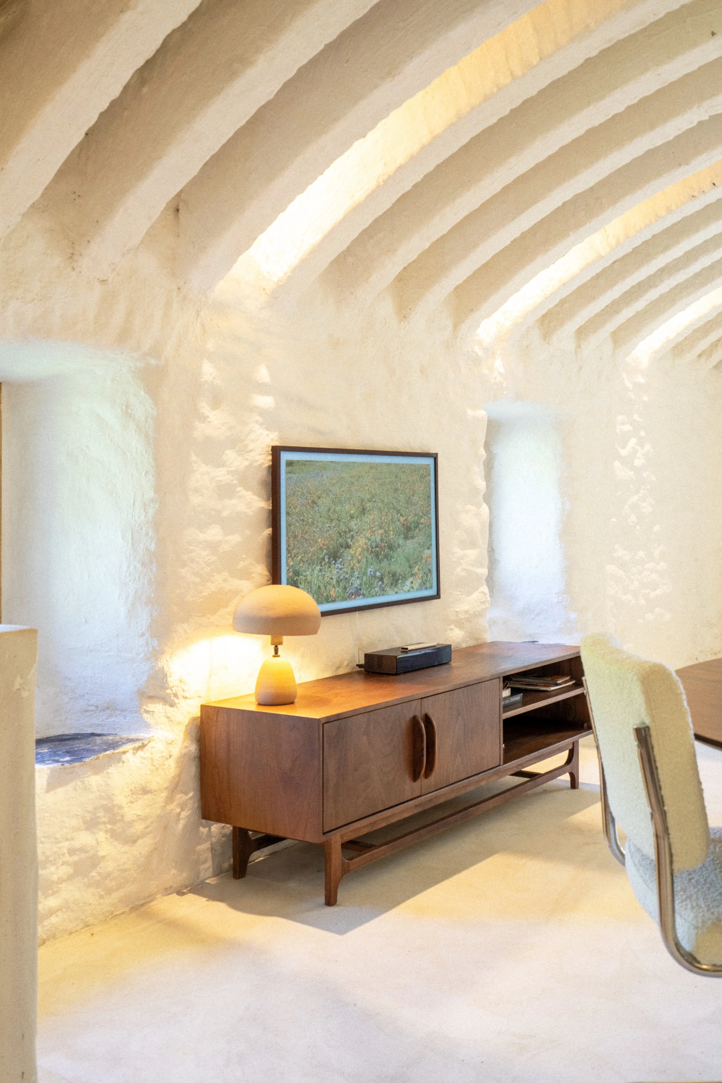 Intérieur d'une pièce avec murs en plâtre blanc et plafond en voûtes, meuble en bois avec une lampe beige, une peinture dans un cadre noir, une télévision et quelques livres, chaise beige à côté d'une table en bois.