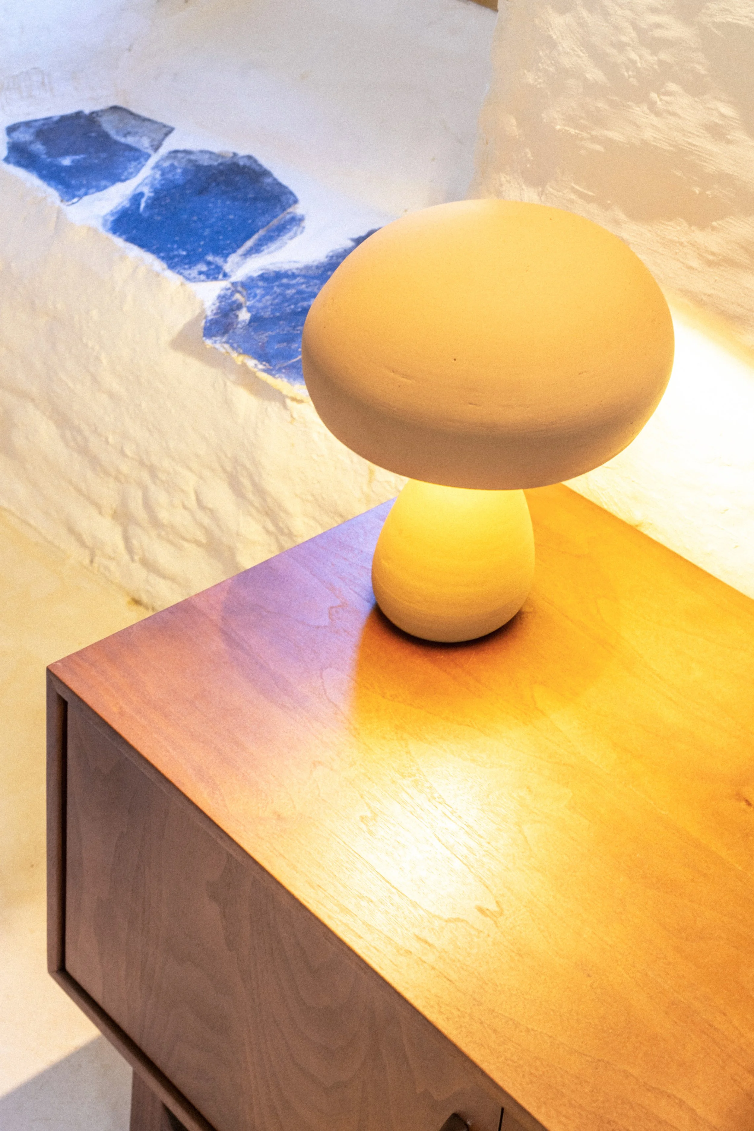 Lampe de table arrondie de couleur beige, posée sur une table en bois clair, dans une pièce avec mur en pierre blanche et une fenêtre avec petites pierres bleues.
