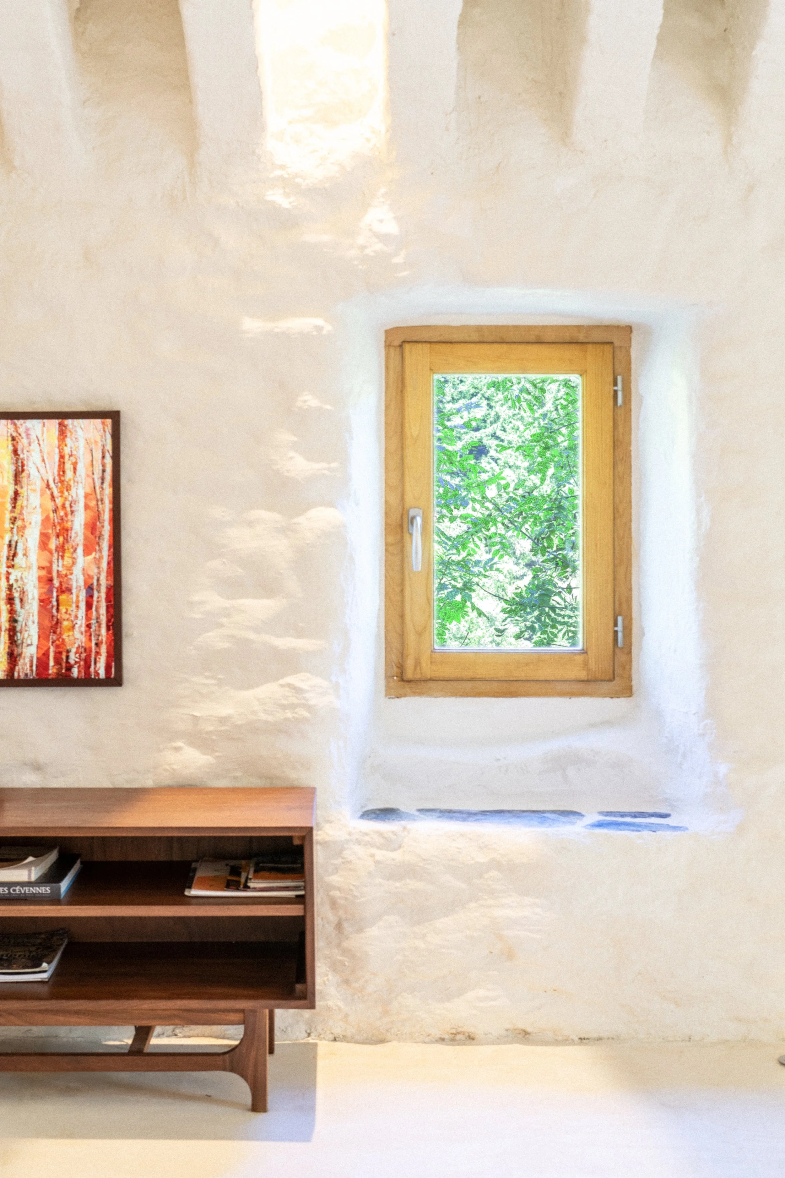 Fenêtre en bois avec vue sur des feuilles vertes, mur blanc rustique avec étagère en bois et une peinture d'arbres rouges.