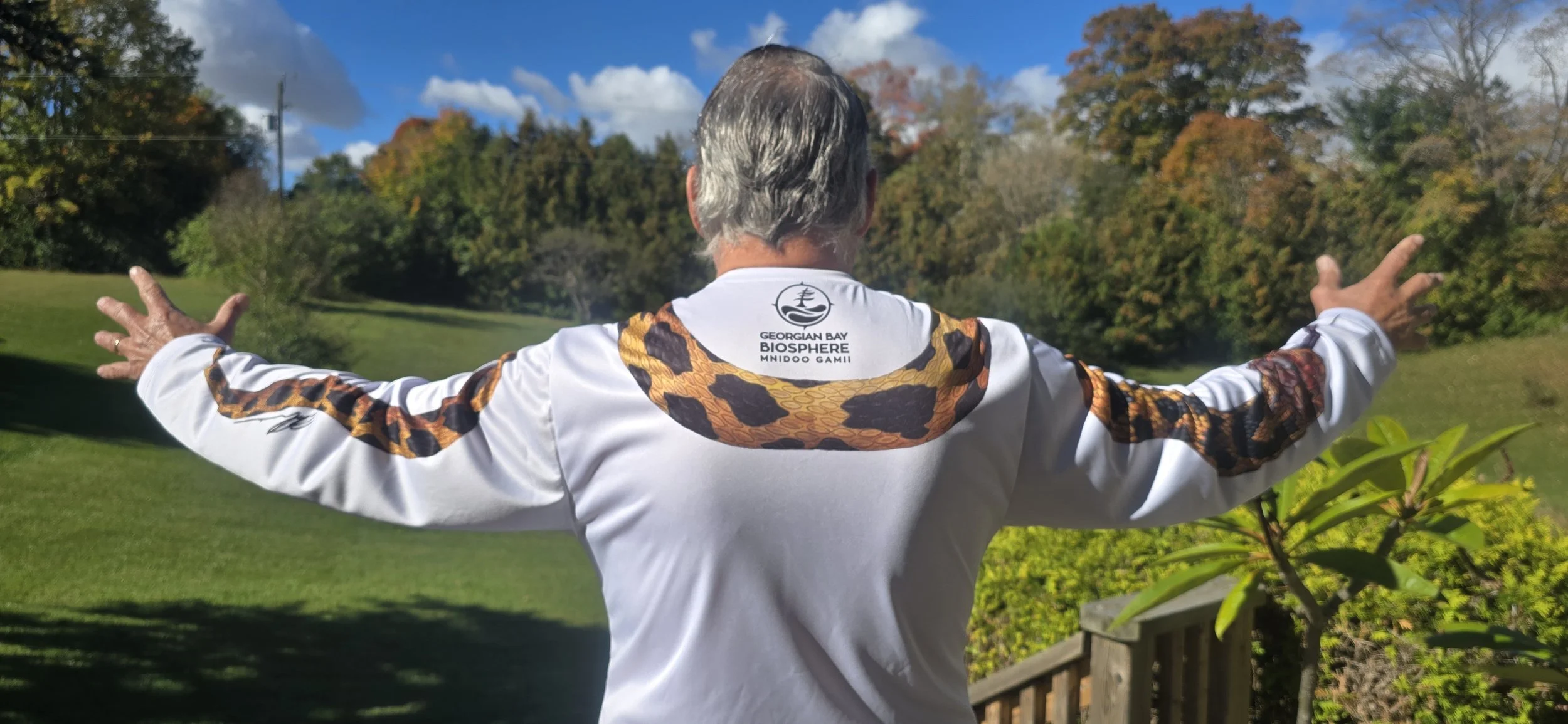 Bert Liverance in a white Georgian Bay Biosphere shirt standing outdoors with arms outstretched in a landscaped garden.