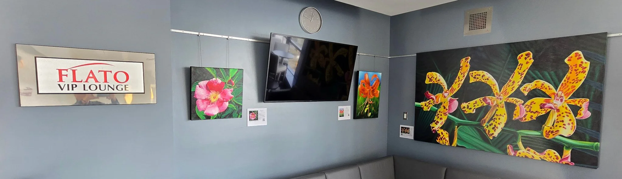 Flato VIP Lounge interior with colorful floral artwork, a wall-mounted TV, clock, and seating, paintings by Bert Liverance