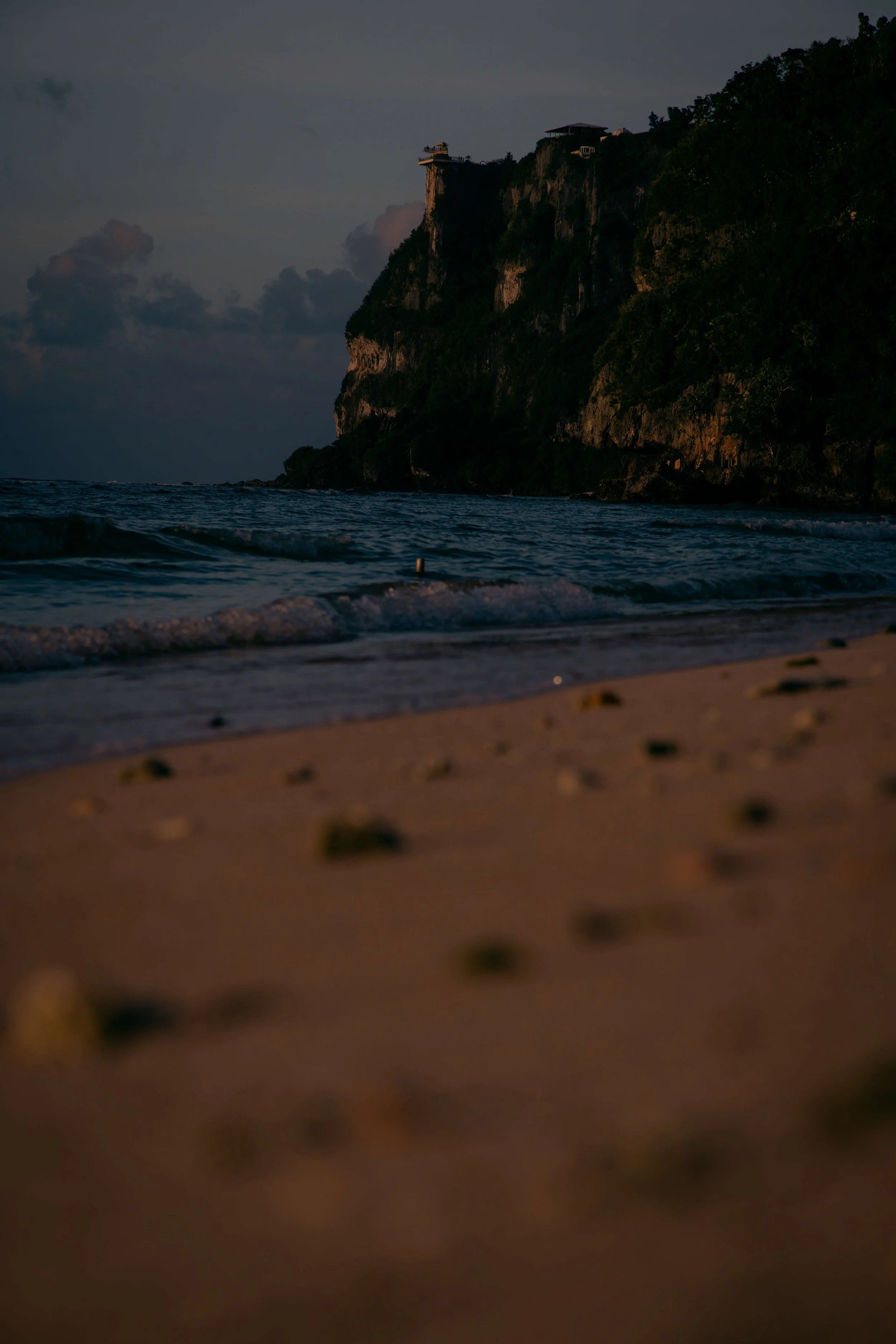 A view of a cliffside with trees and buildings, overlooking the ocean during dusk or dawn, with dark clouds in the sky.