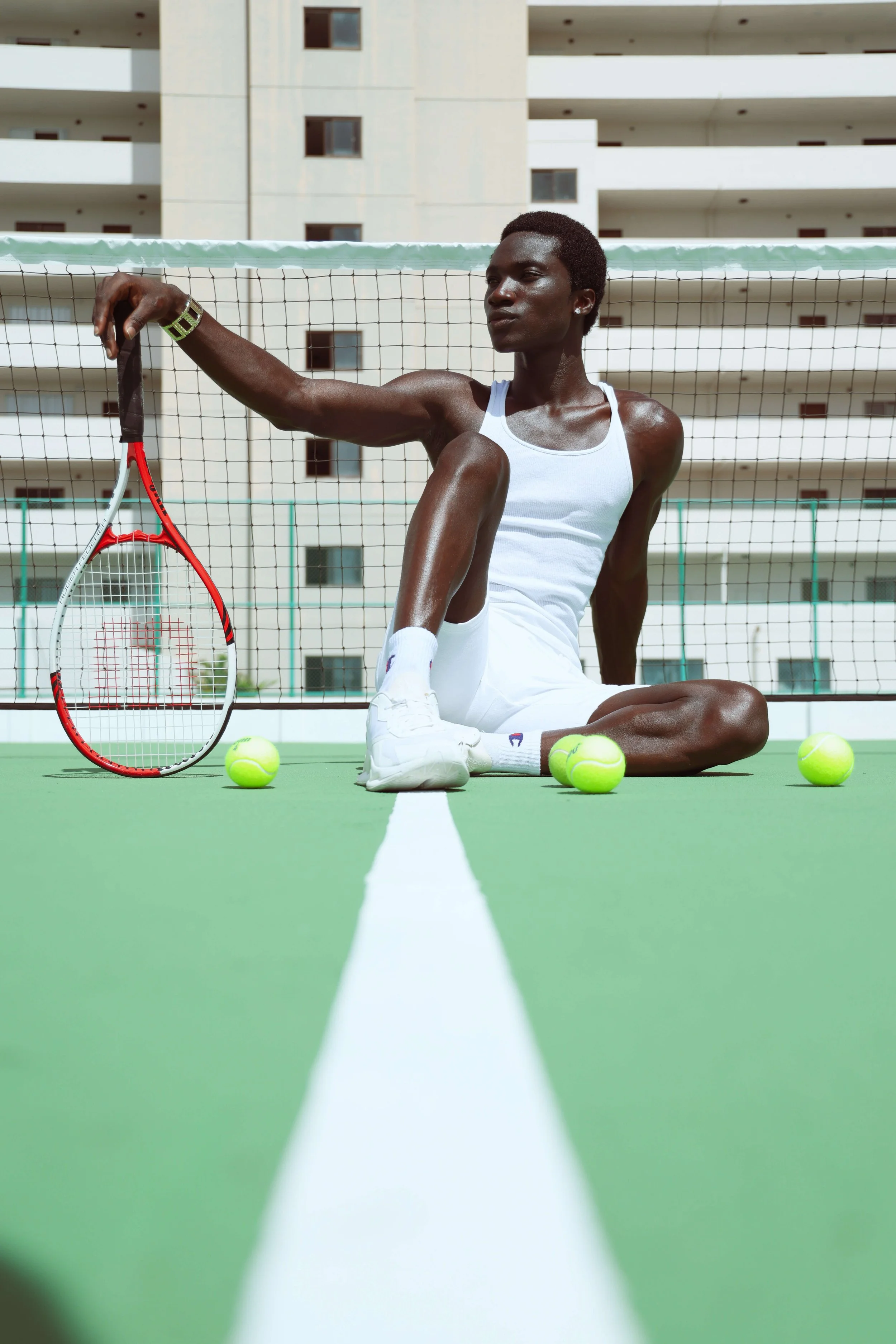 A person sitting on a tennis court with tennis balls and a tennis racket, against a background of a building with balconies.