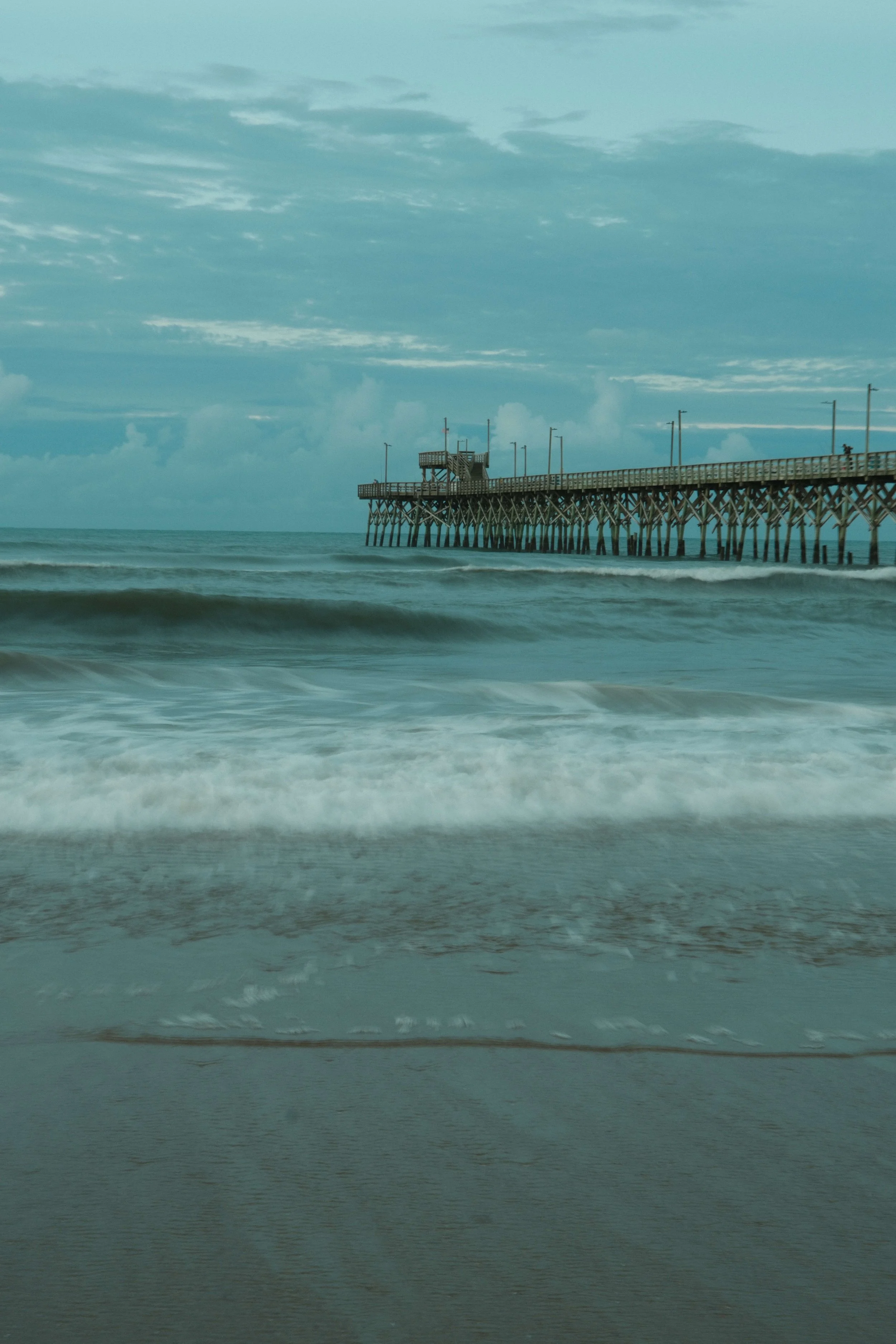 Long pier extending into the ocean with a watchtower at the end, cloudy sky above, waves crashing on the shore.