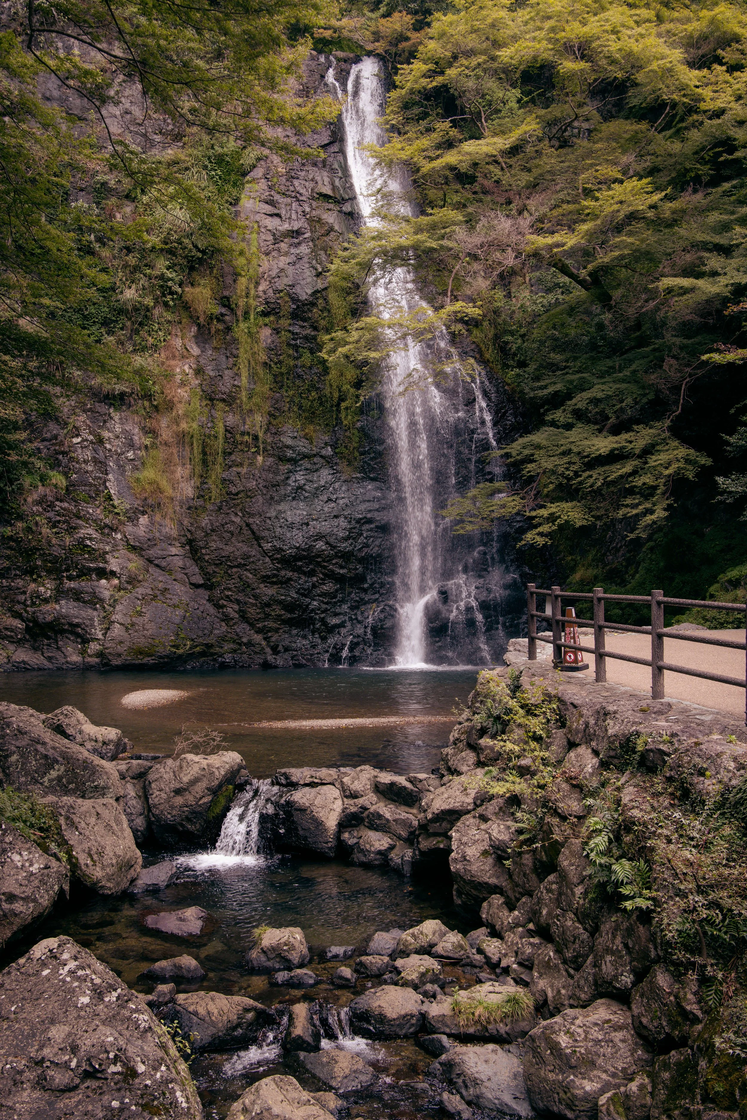 A waterfall flowing down a rocky cliff surrounded by lush green trees, with a small water pool at the base and a wooden railing on the right side of the image.