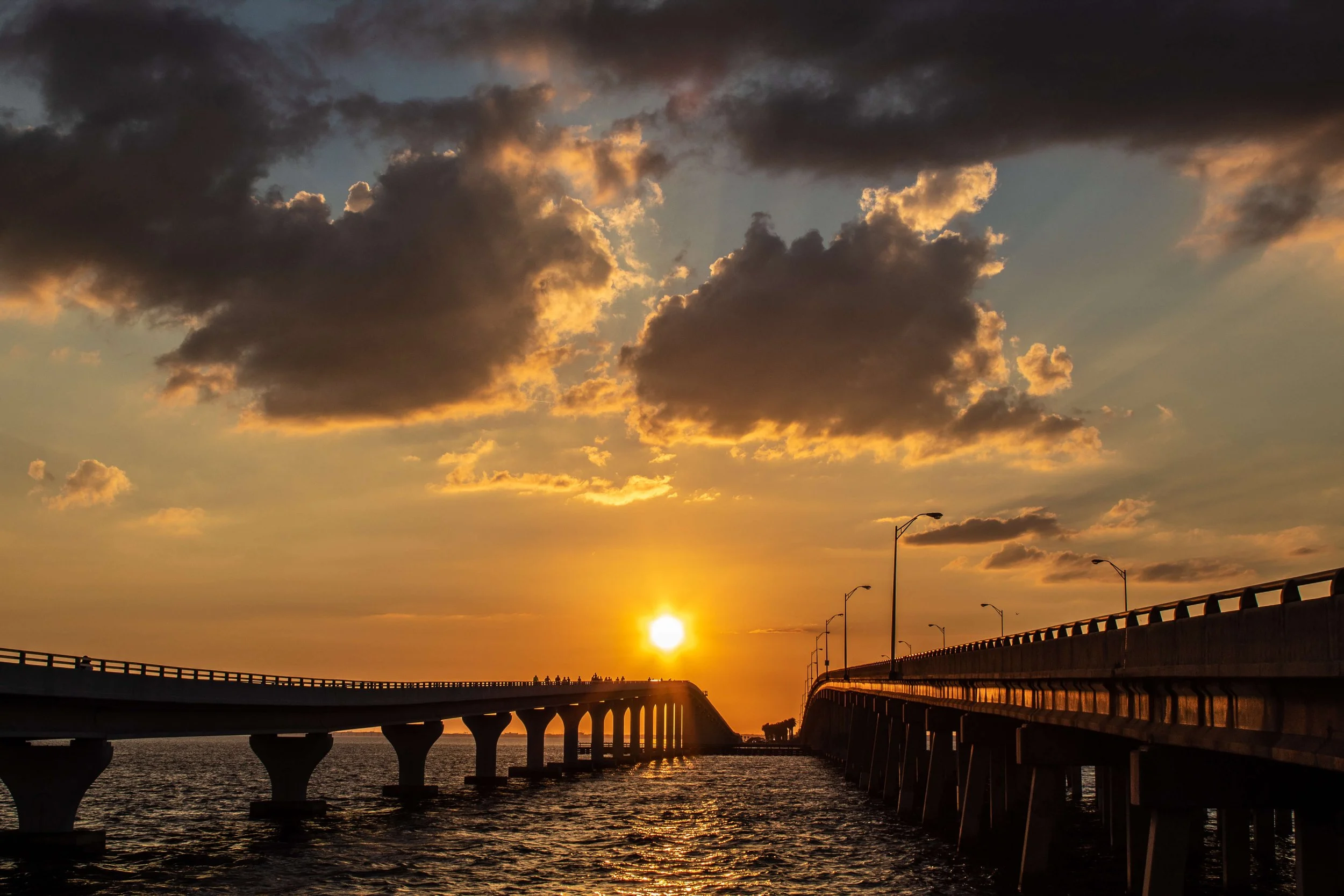 Sunset behind a bridge over a body of water with cloudy sky and streetlights along the bridge
