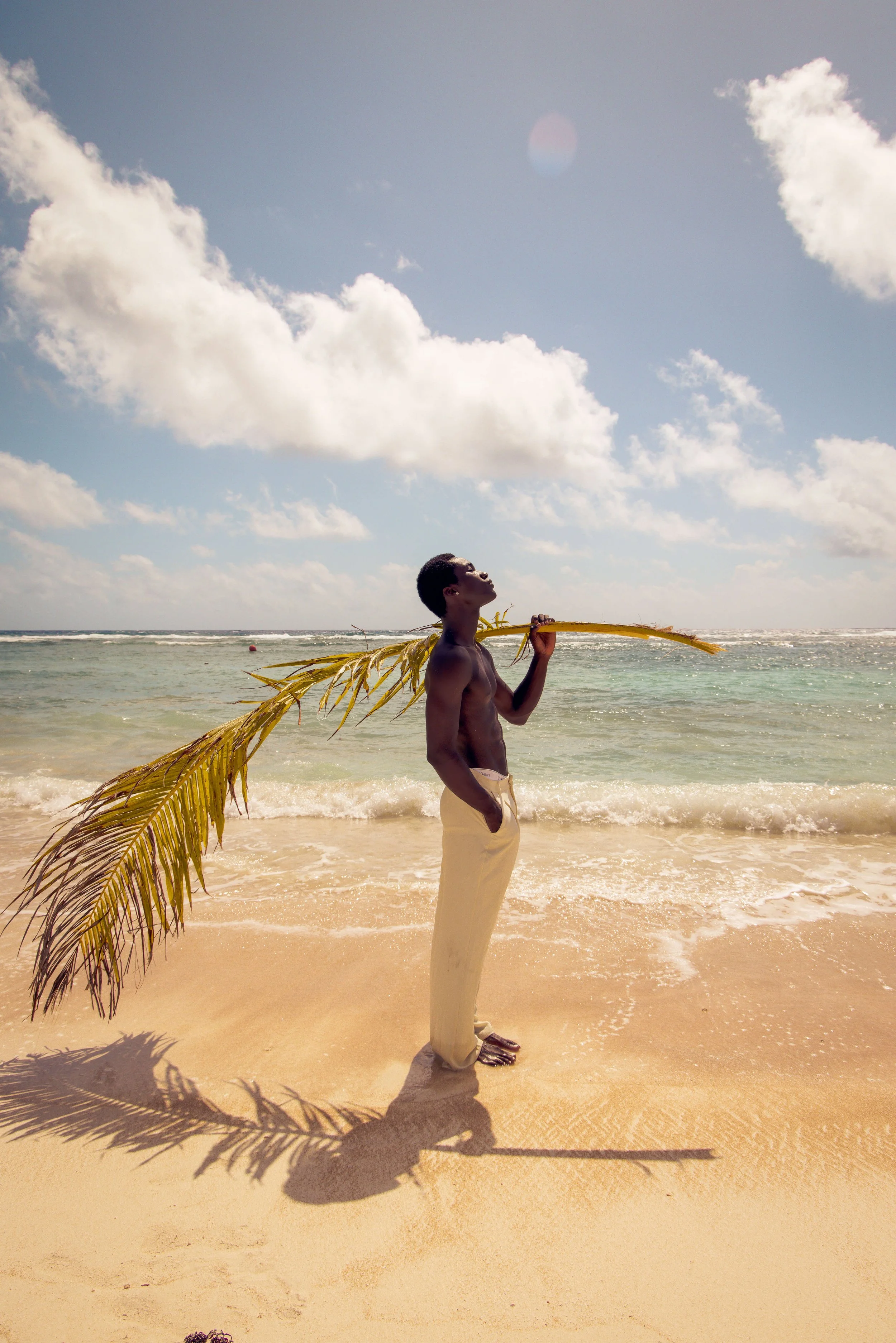 A young man standing on a sandy beach holding a yellow palm frond over his shoulder, with the shadow of the palm cast on the sand.