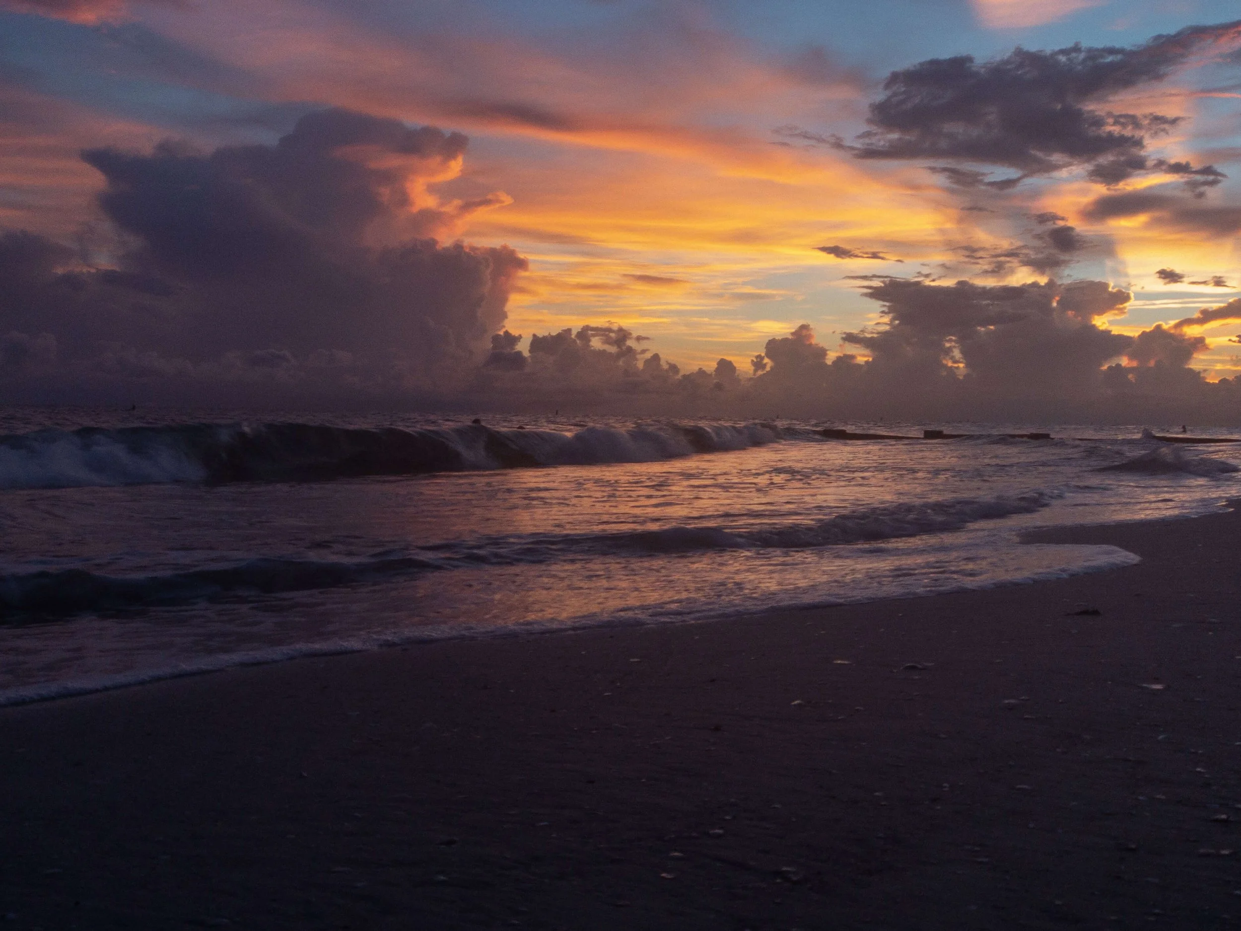 Sunset over the ocean with vibrant orange, pink, purple, and blue hues in the sky, and waves crashing on the sandy beach in the foreground.