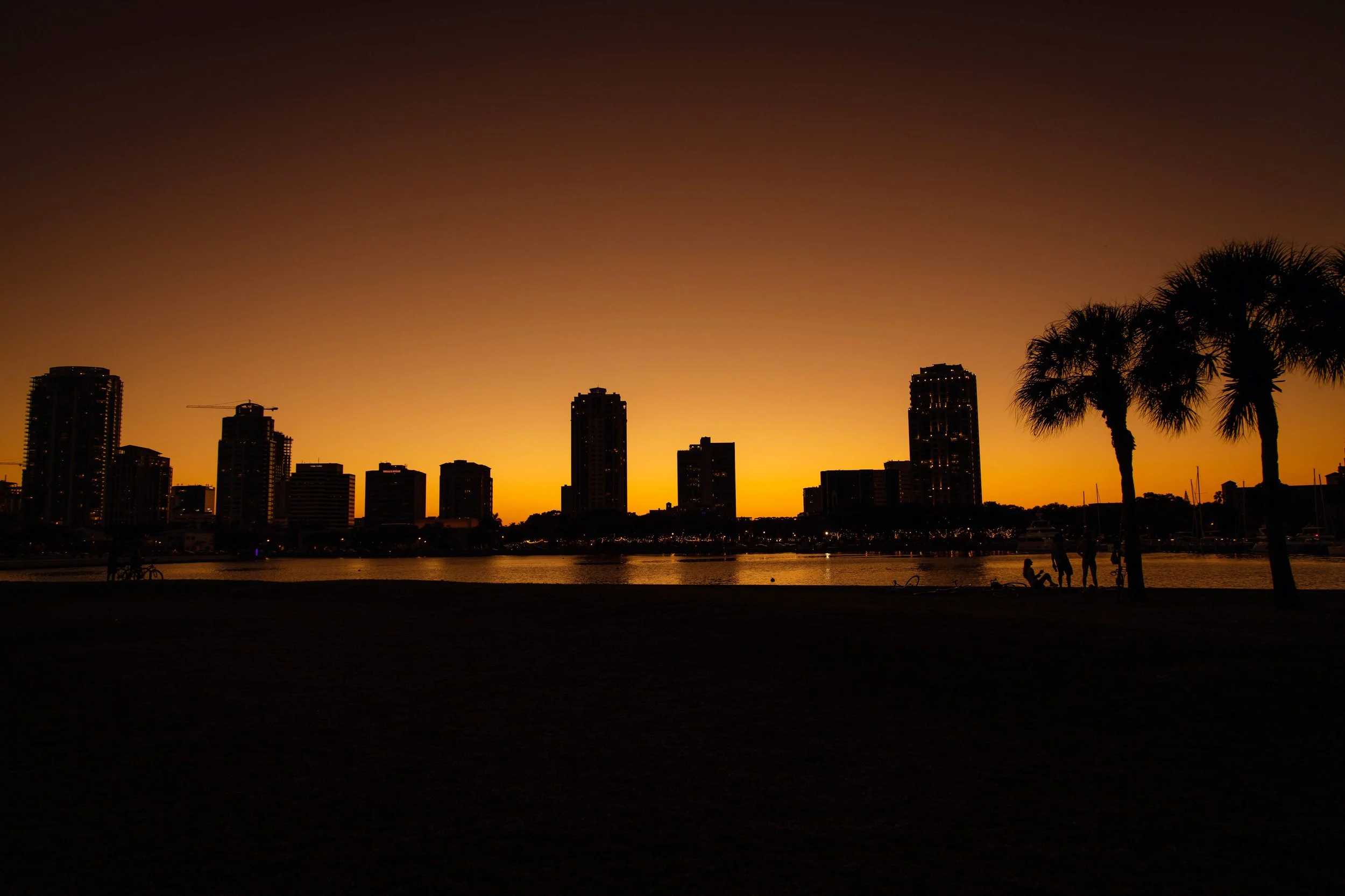 Skyline of tall buildings during sunset with a dark foreground, silhouetted palm trees, and people along the water's edge.
