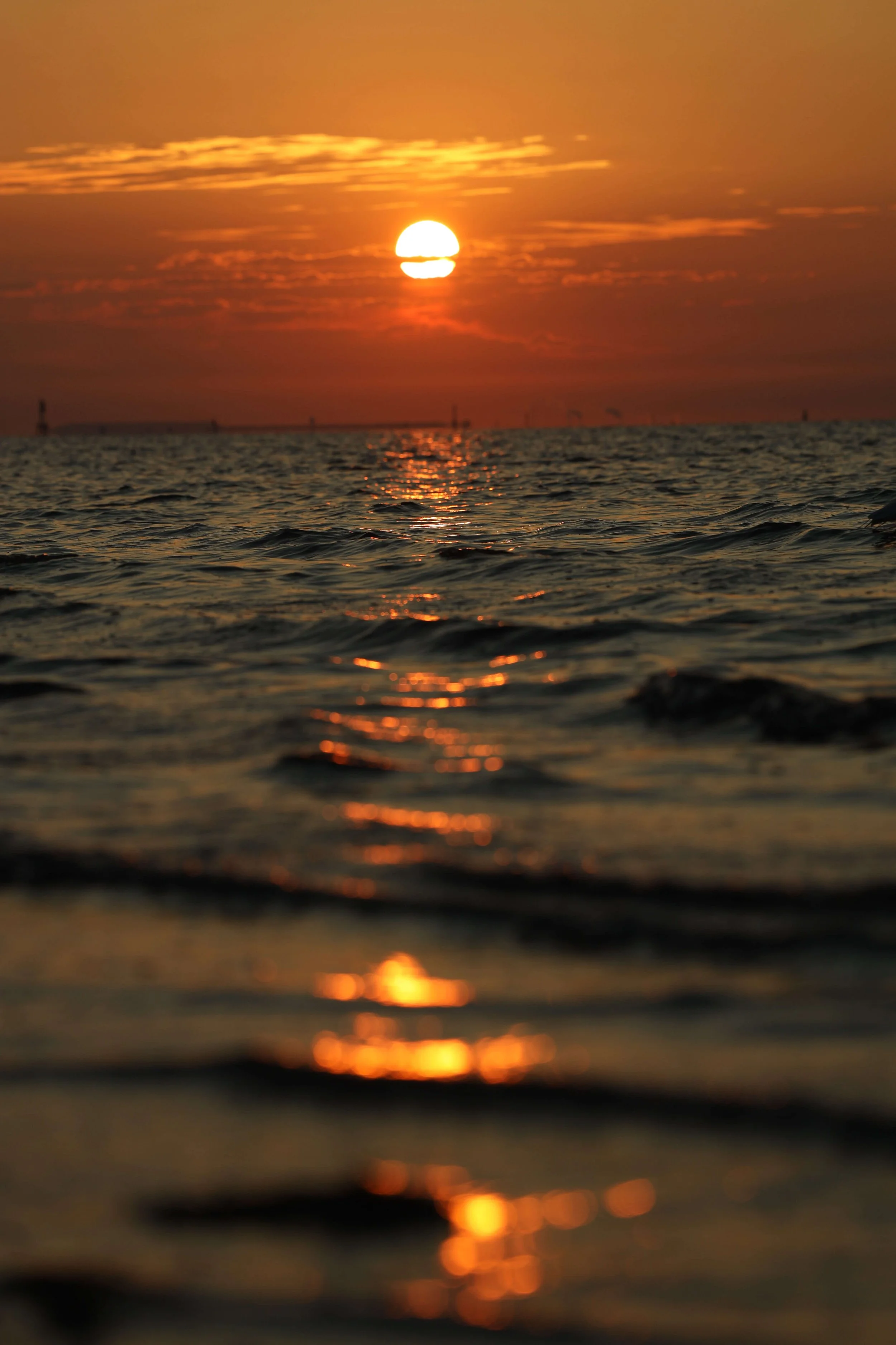 Sunset over the ocean with the sun partially obscured by clouds, reflecting a golden glow on the water's surface.