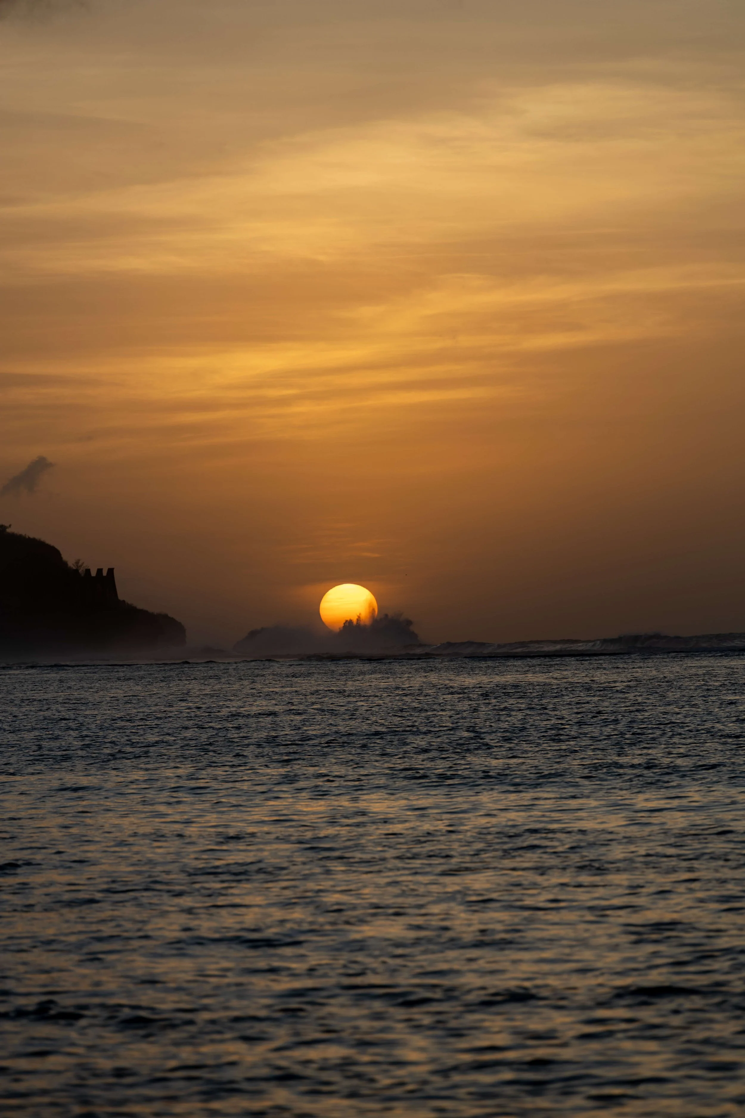 Sunset over the ocean with a silhouette of a cliff in the foreground and cloudy sky.
