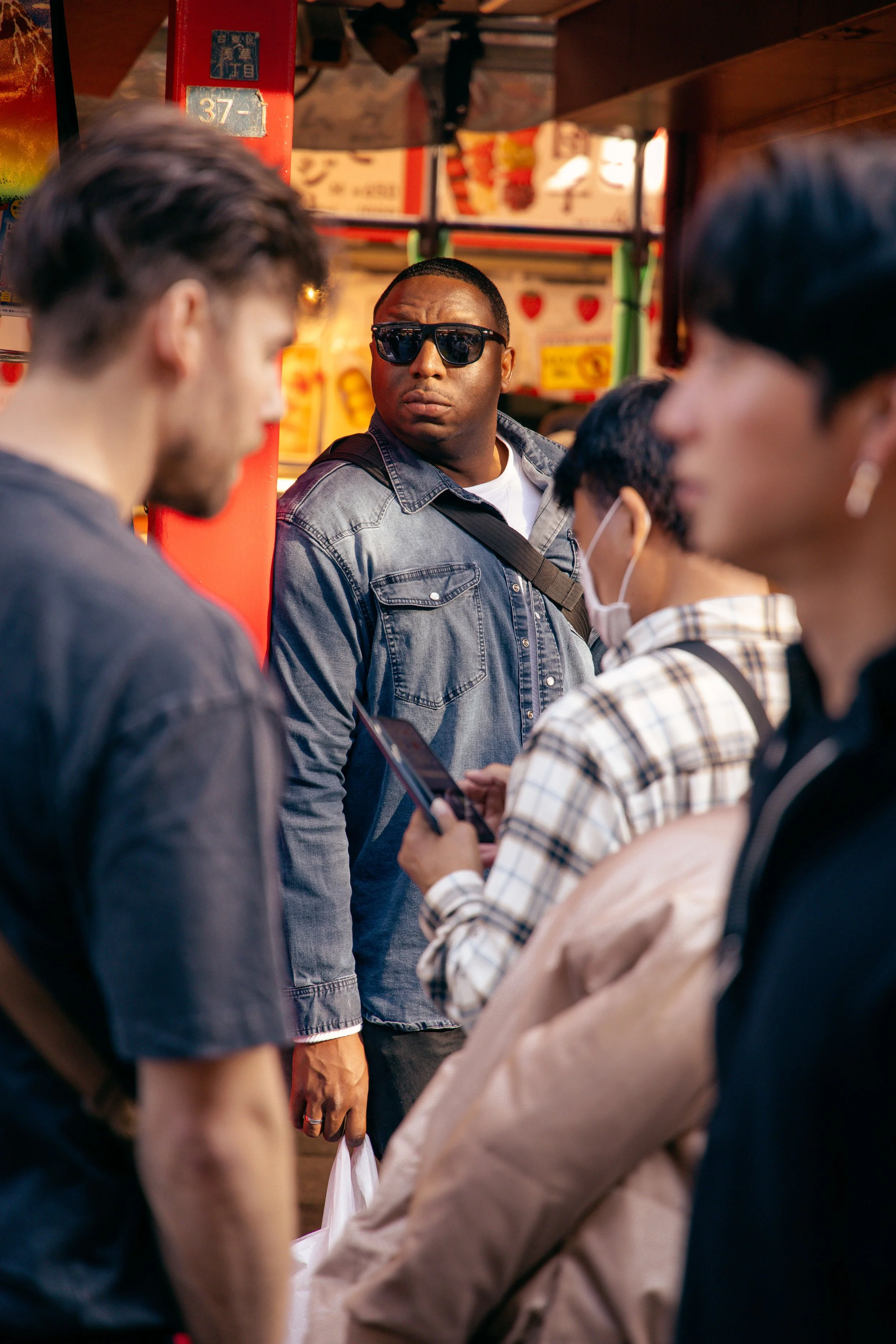 A man wearing sunglasses and a denim jacket looking at three women in an outdoor market, with colorful signs and items in the background.