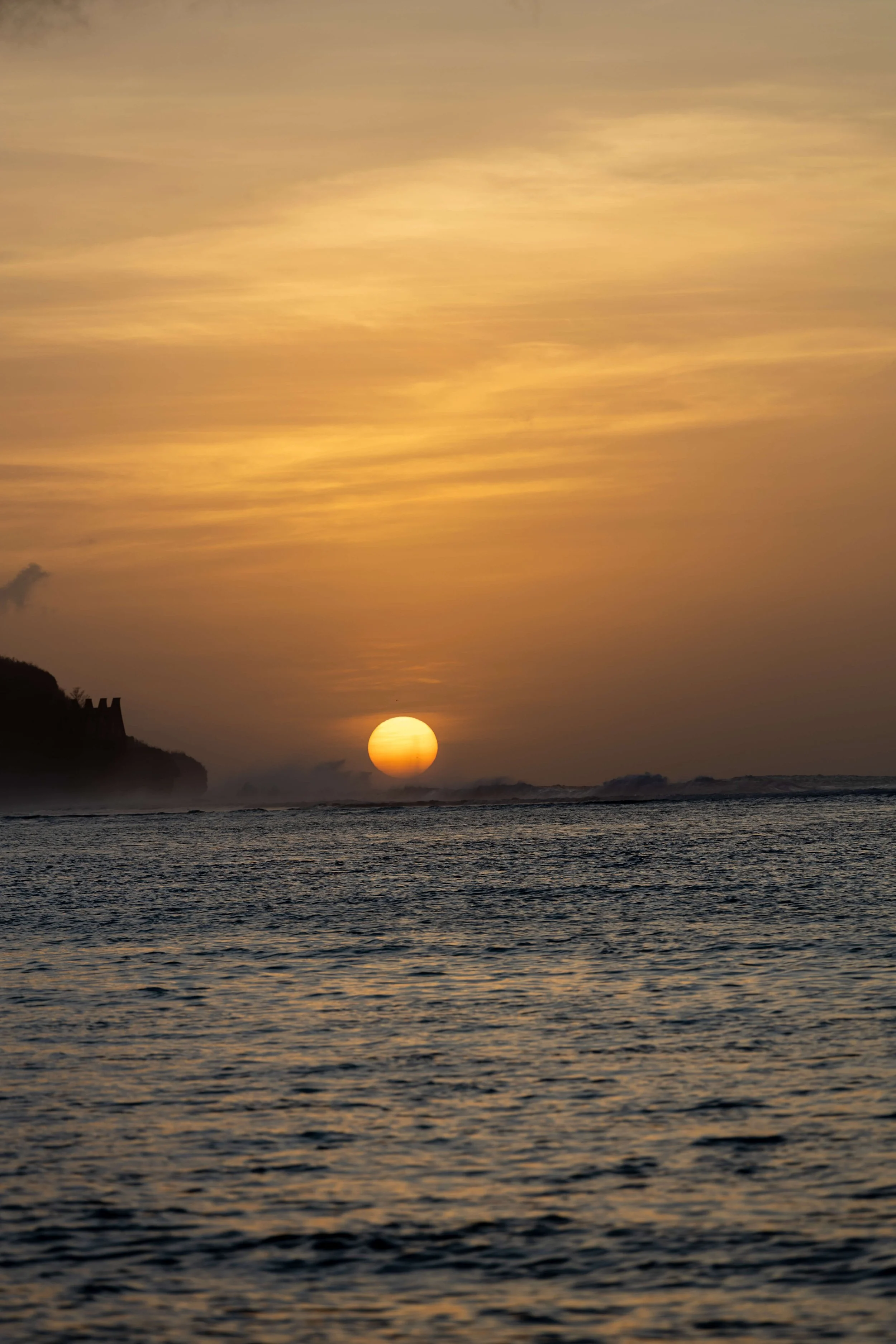 Sunset over the ocean with clouds in the sky and a silhouette of land on the horizon.