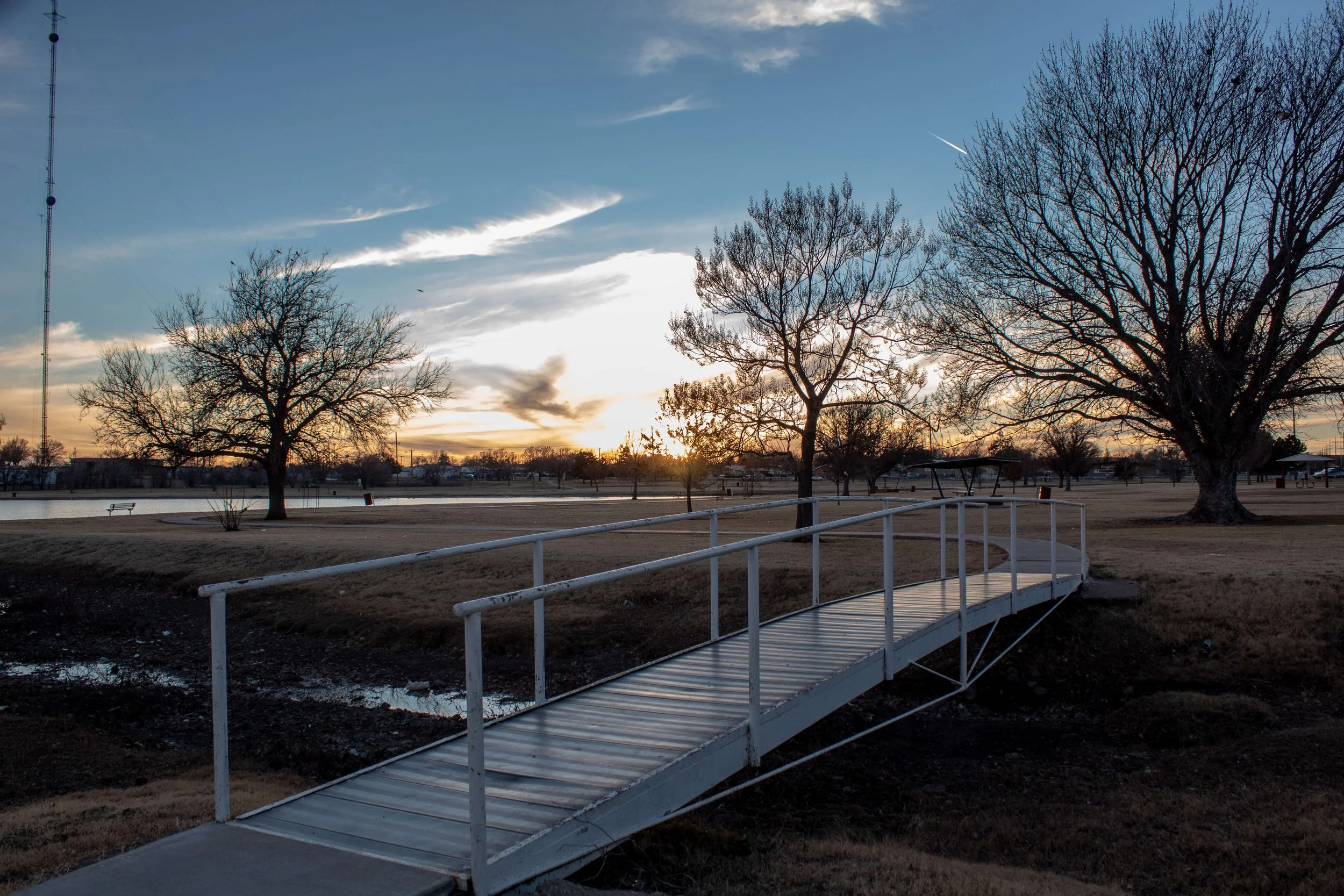 A metal pedestrian bridge over muddy ground near a park with leafless trees and a body of water in the background at sunset.