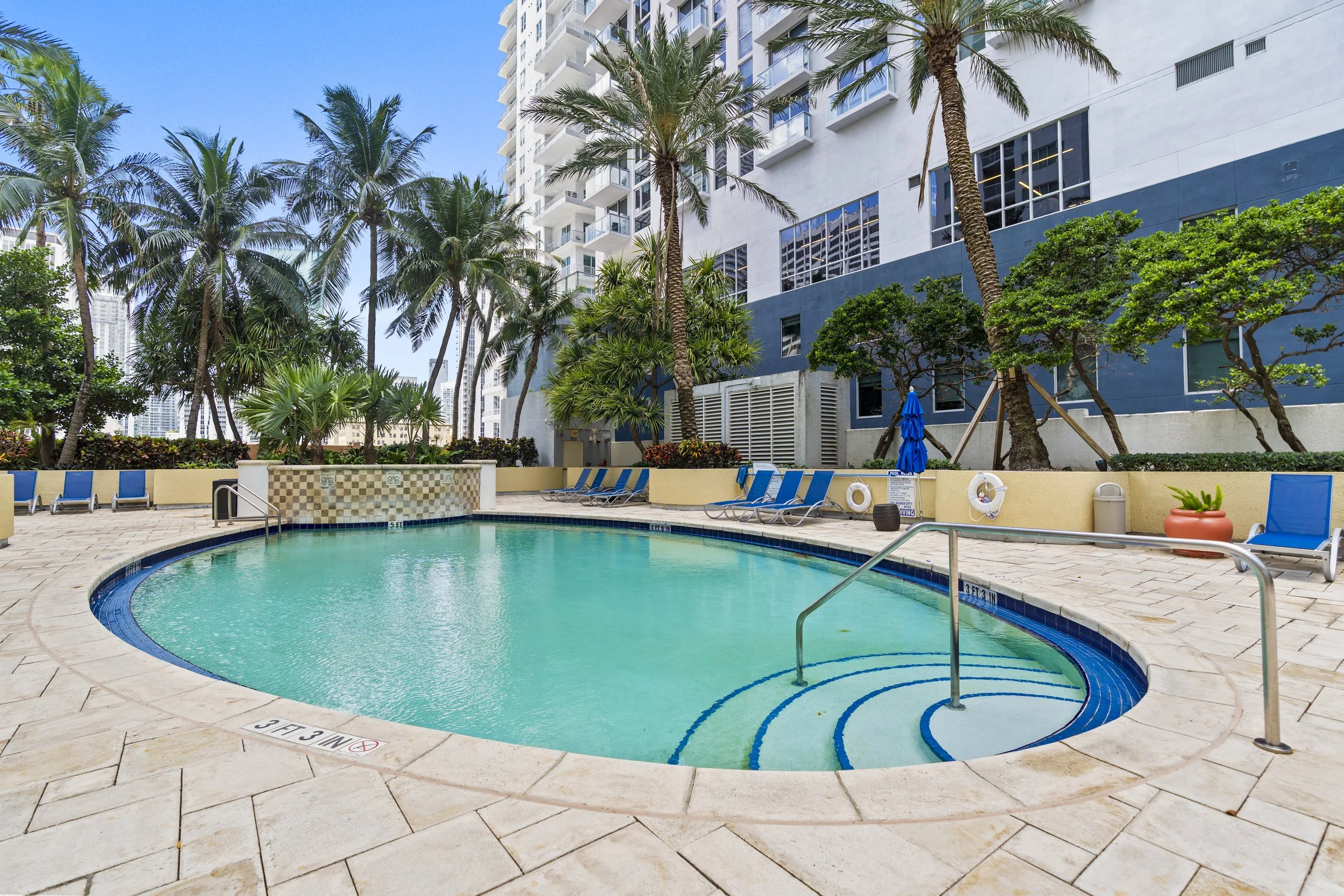 Outdoor swimming pool area surrounded by tall palm trees, lounge chairs, a building with large windows, and a clear blue sky.