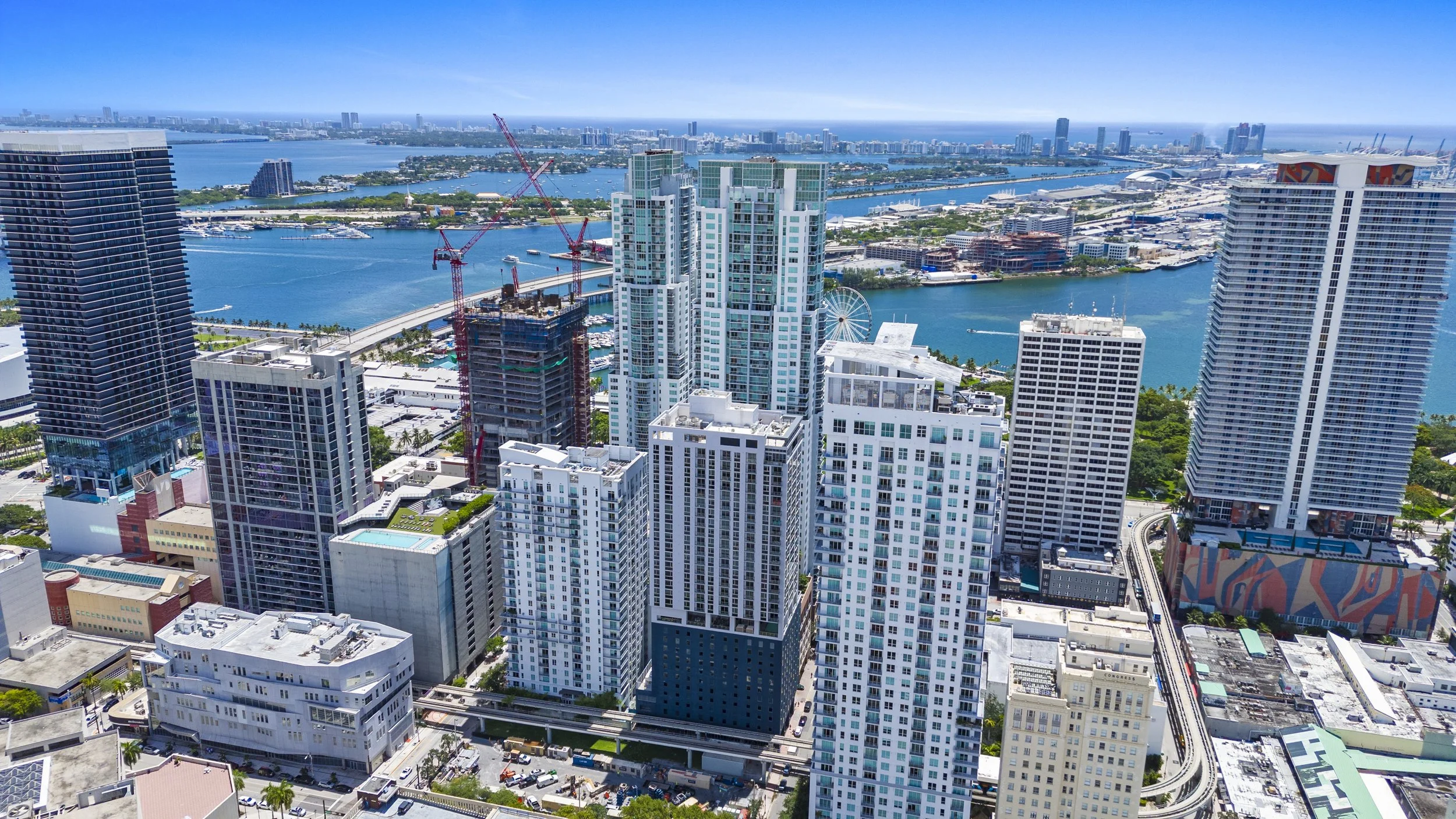 Aerial view of a city skyline with high-rise buildings, a waterway with boats, and a distant ocean under a clear blue sky.
