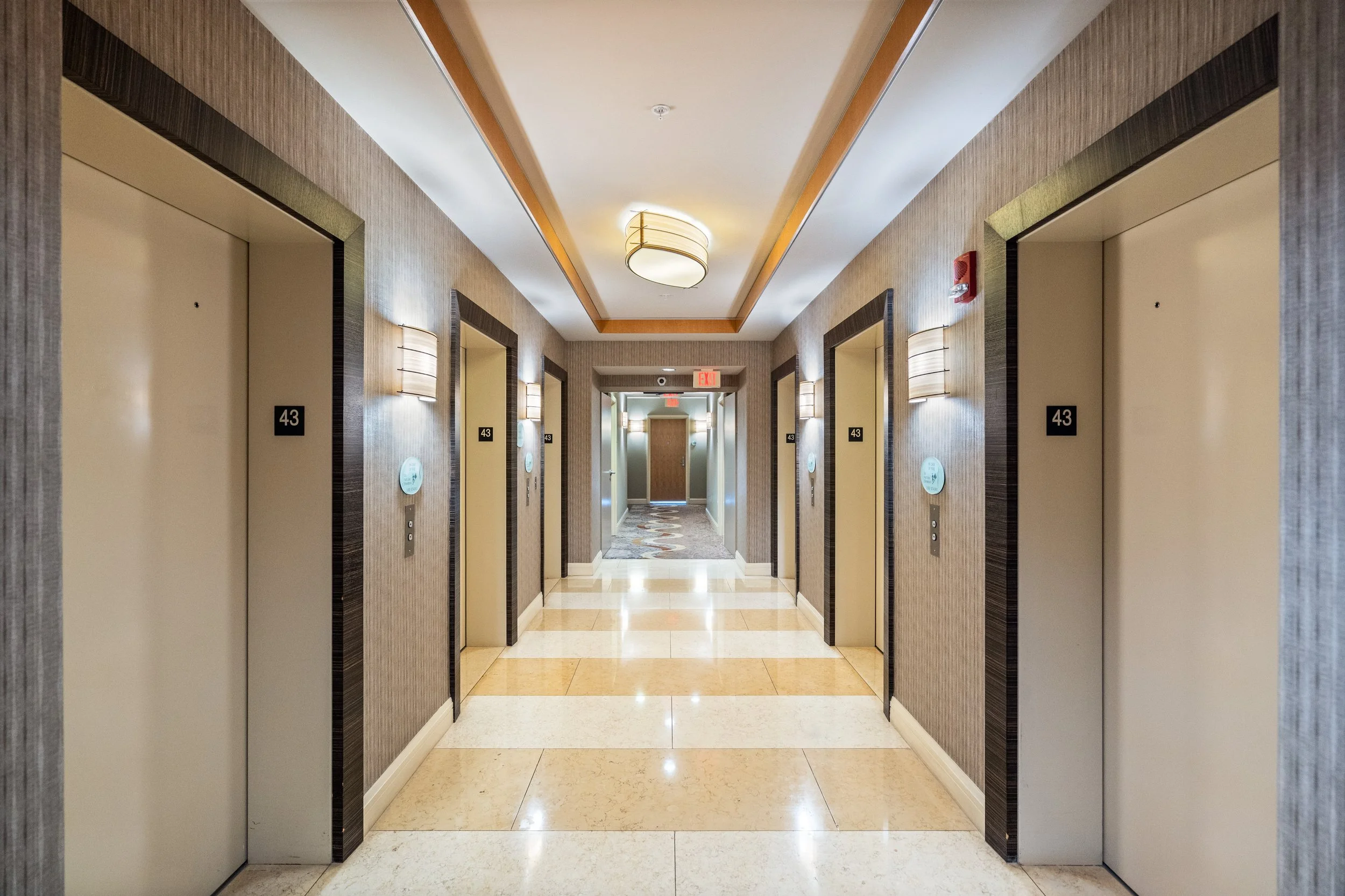 Hotel hallway with elevators on each side, room number 43, ceiling lights, fire alarm, and exit sign at the end of the corridor.