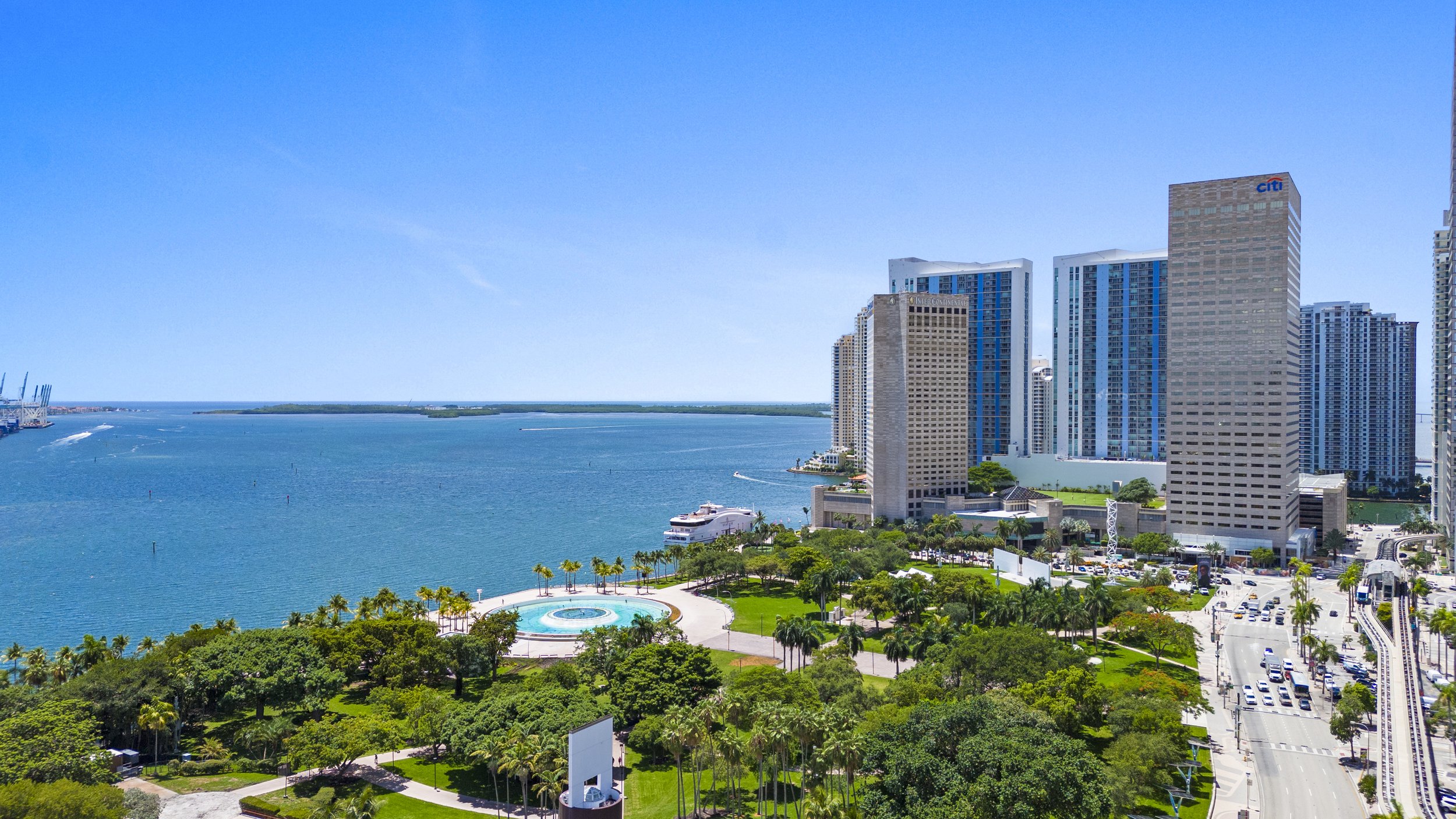 City skyline with high-rise buildings next to water, green park with trees and a fountain, clear blue sky.