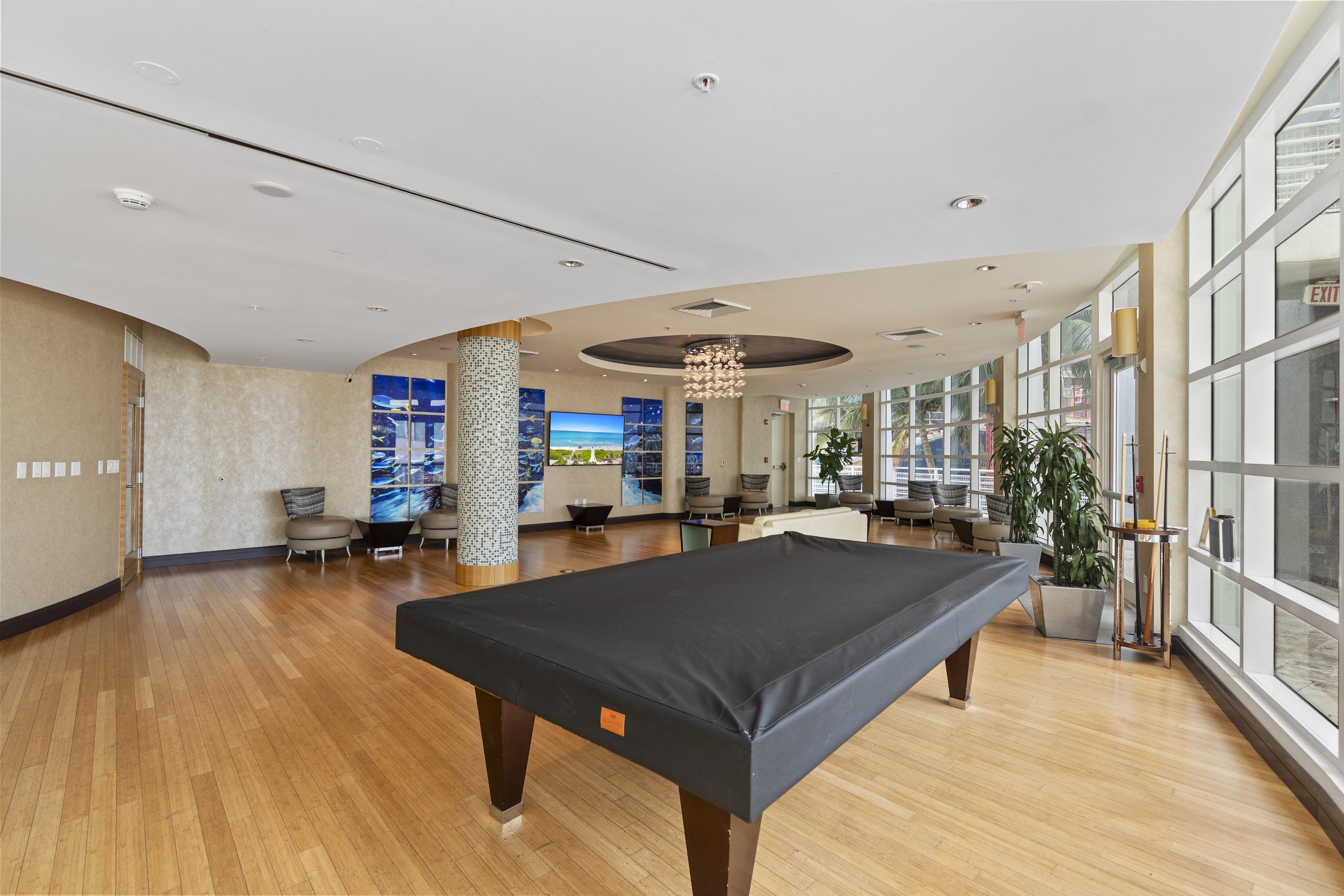 An indoor lounge area with large windows on the right side, wooden flooring, a black pool table in the foreground, a cluster of chairs with a small table in the background, a mosaic column, wall-mounted screens displaying a beach scene, and a modern 