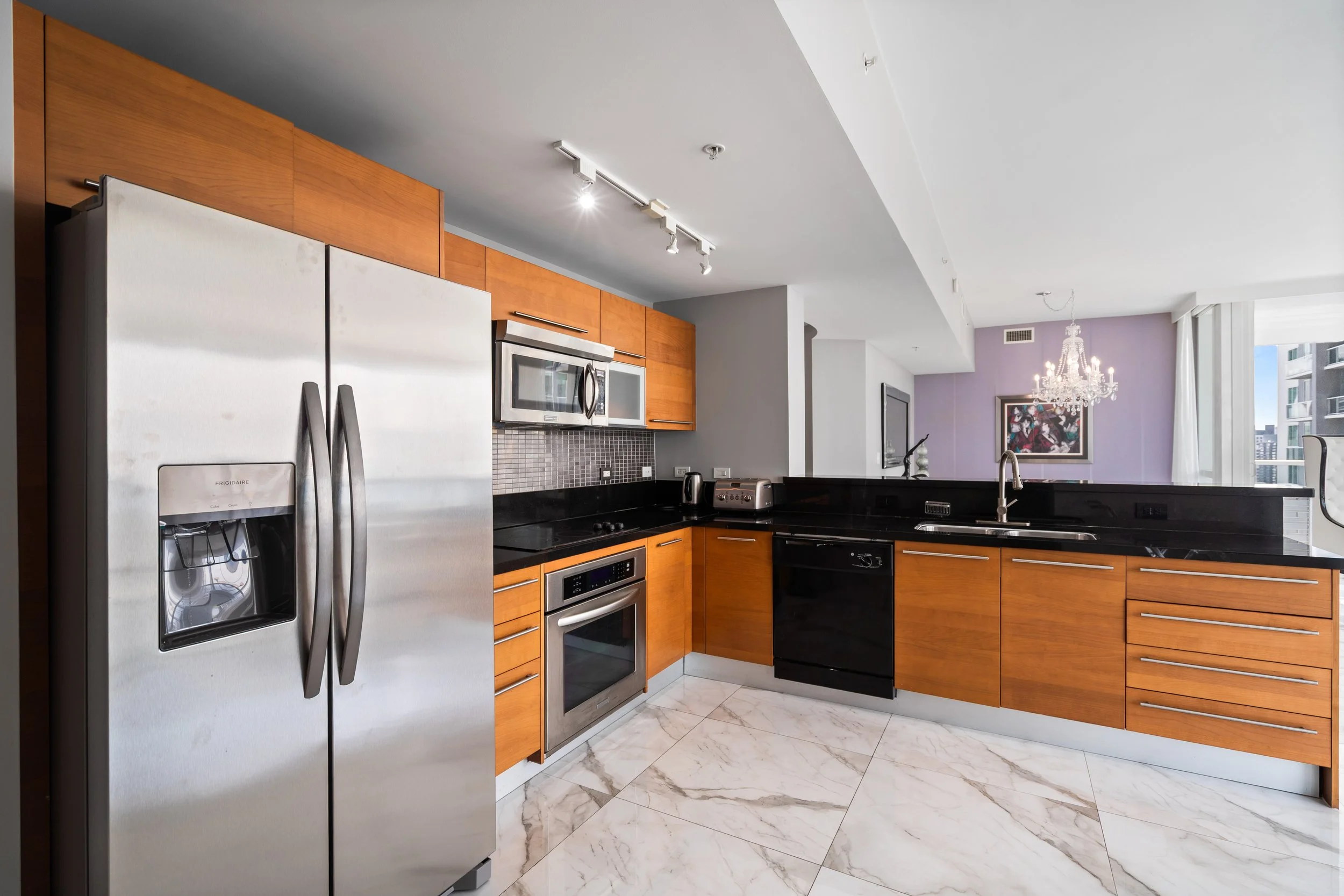 Modern kitchen with wooden cabinets, stainless steel refrigerator, microwave, oven, black countertop, and white marble floor. A living area with a purple wall, a chandelier, and large windows is visible in the background.