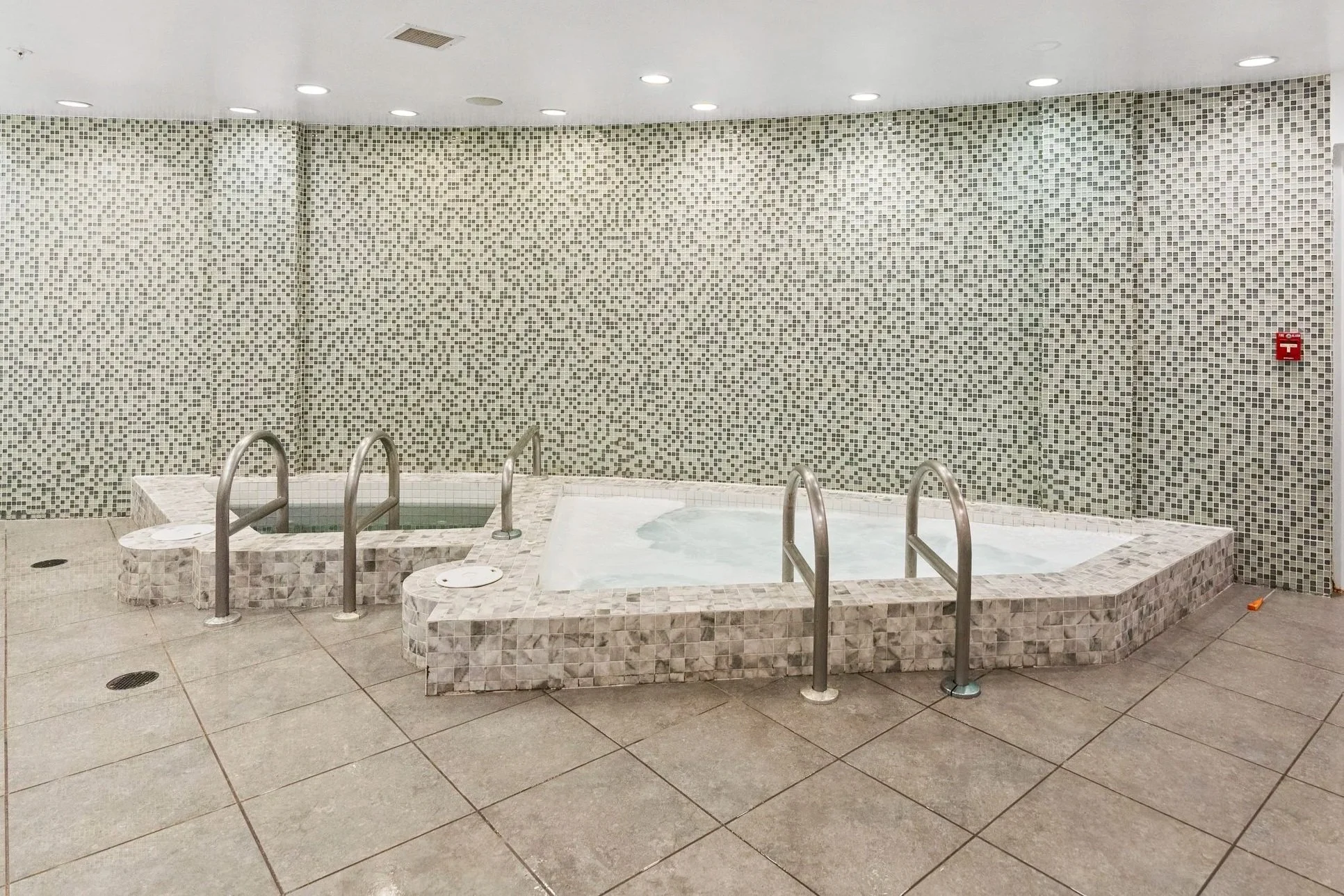Indoor spa area with a hot tub and a smaller whirlpool pulled by stainless steel railings, surrounded by beige and gray tiles and a mosaic wall, with a red fire alarm on the wall.