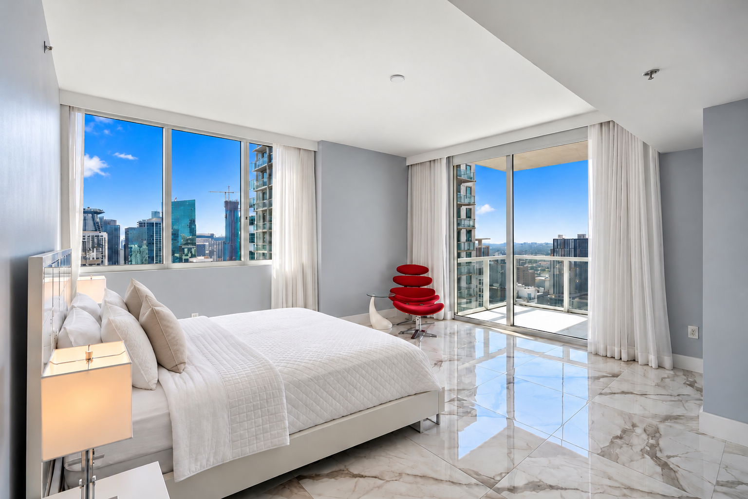 Modern bedroom with large windows showing cityscape, white bed with beige pillows, side table with lamp, red ergonomic chair near glass balcony door, marble floor, white curtains.