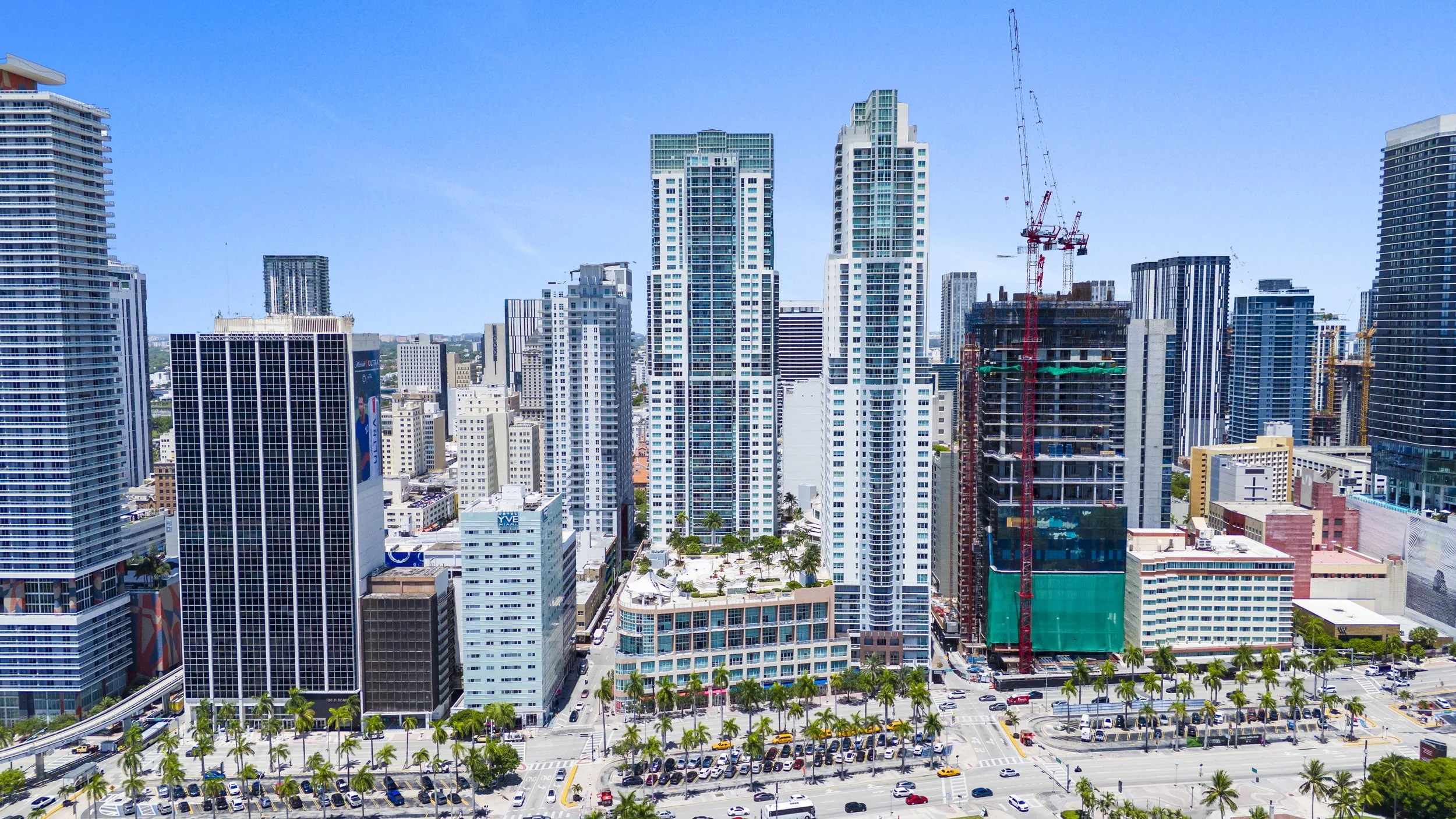 A city skyline with numerous tall modern buildings, some under construction with cranes, and a parking lot filled with cars and palm trees in the foreground.