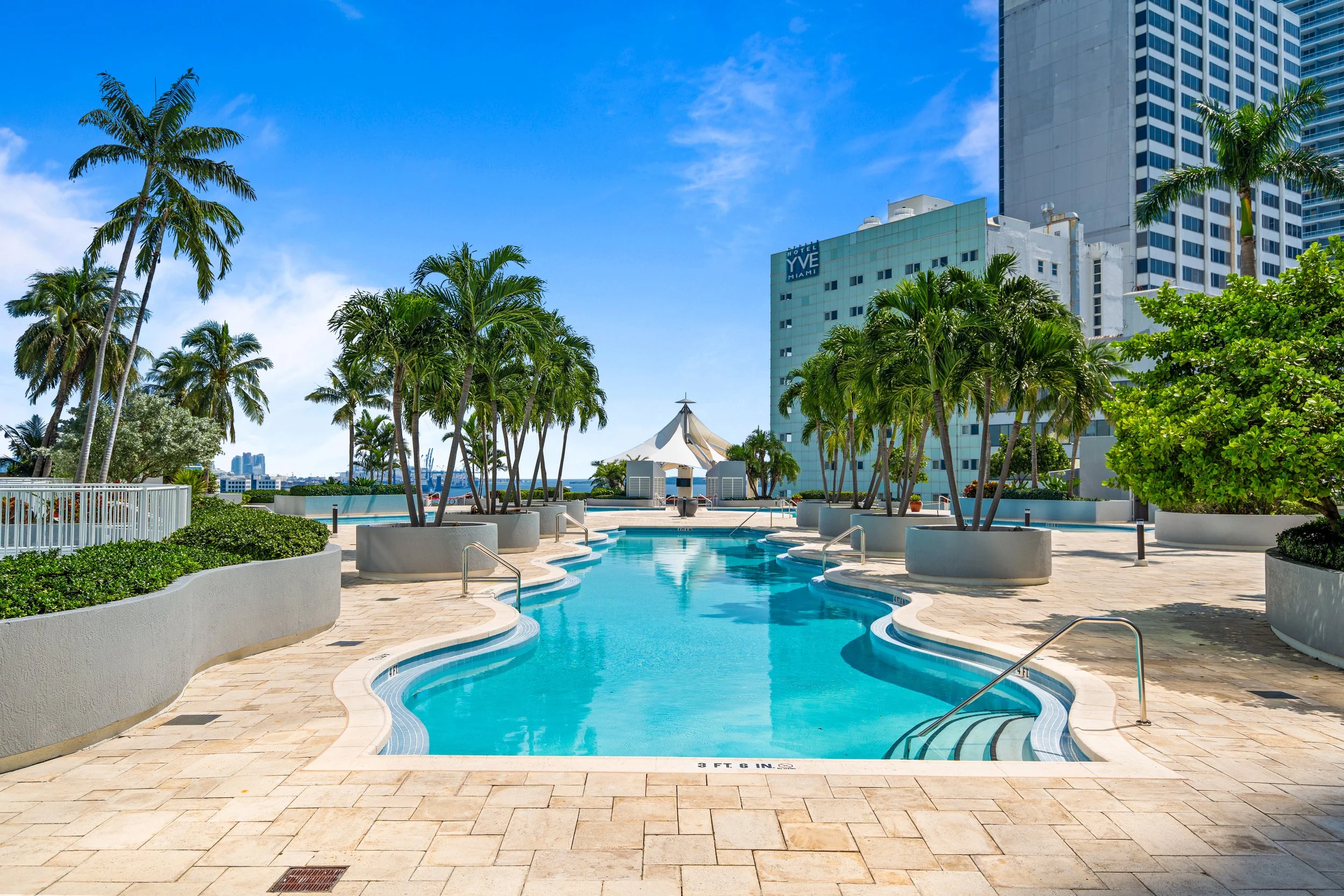 Outdoor swimming pool area surrounded by palm trees, with tall buildings in the background on a clear, sunny day in Miami.