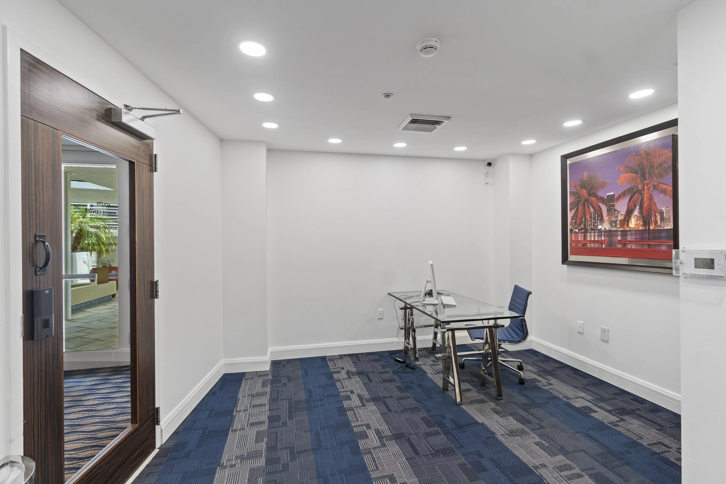 An empty meeting room with white walls, dark blue carpet, a glass conference table, and three chairs. A large framed cityscape picture of a tropical city with palm trees is on the wall. The room has a door with glass and wood trim, and ceiling lights