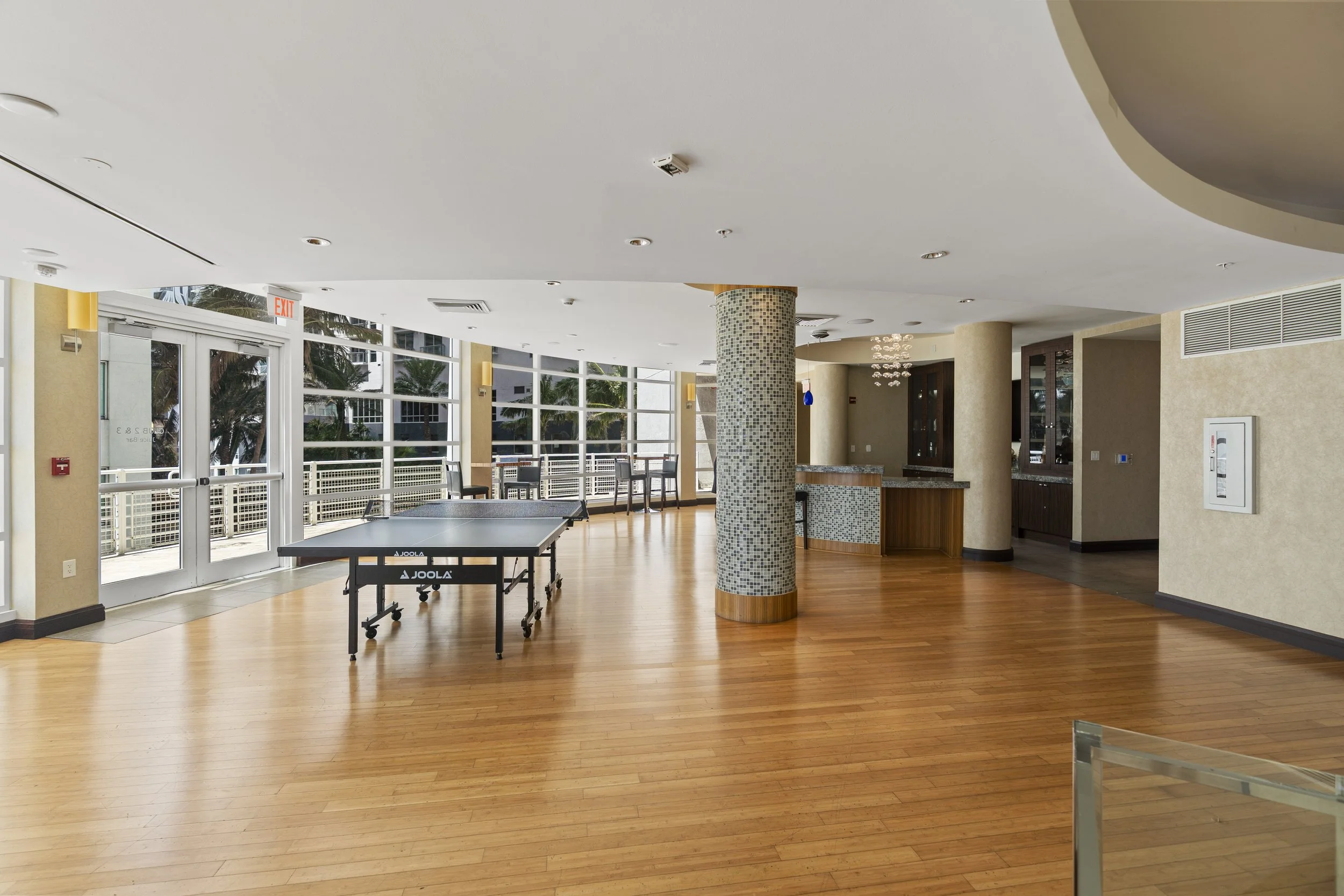 An indoor recreational area with wooden floors, large windows, and a table tennis table. There is a mosaic-covered column in the center and a seating area with chairs and a bar in the background. Natural light streams in through the windows, showing 