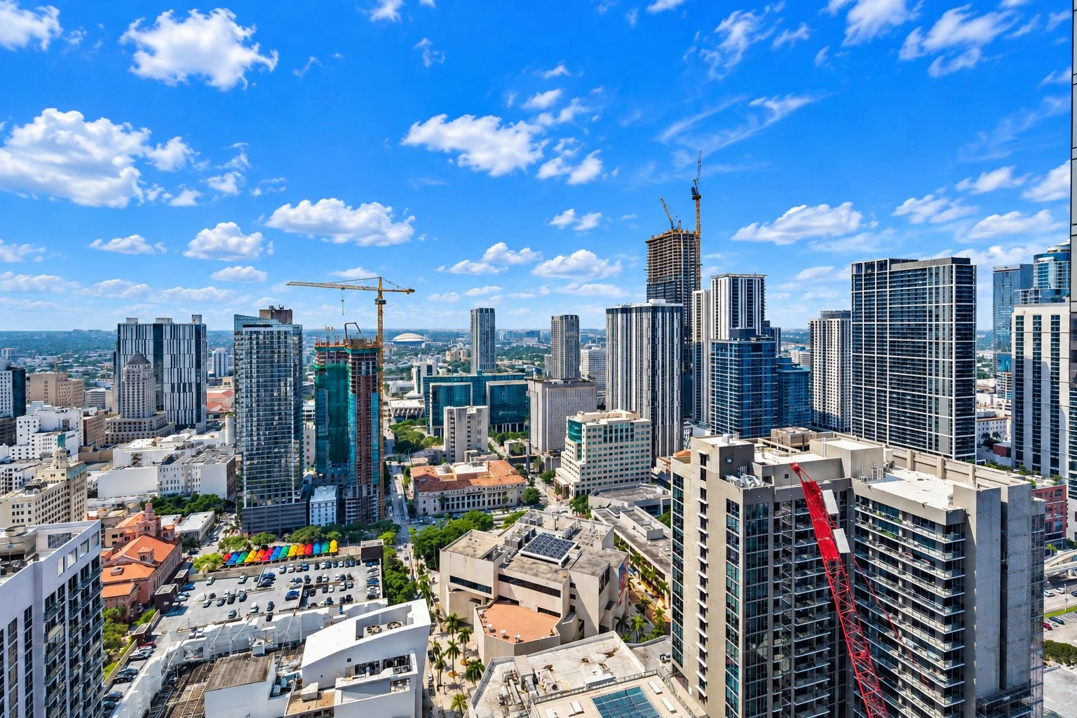 A city skyline with tall modern buildings under a bright blue sky with scattered white clouds. Construction cranes are visible among the high-rises.