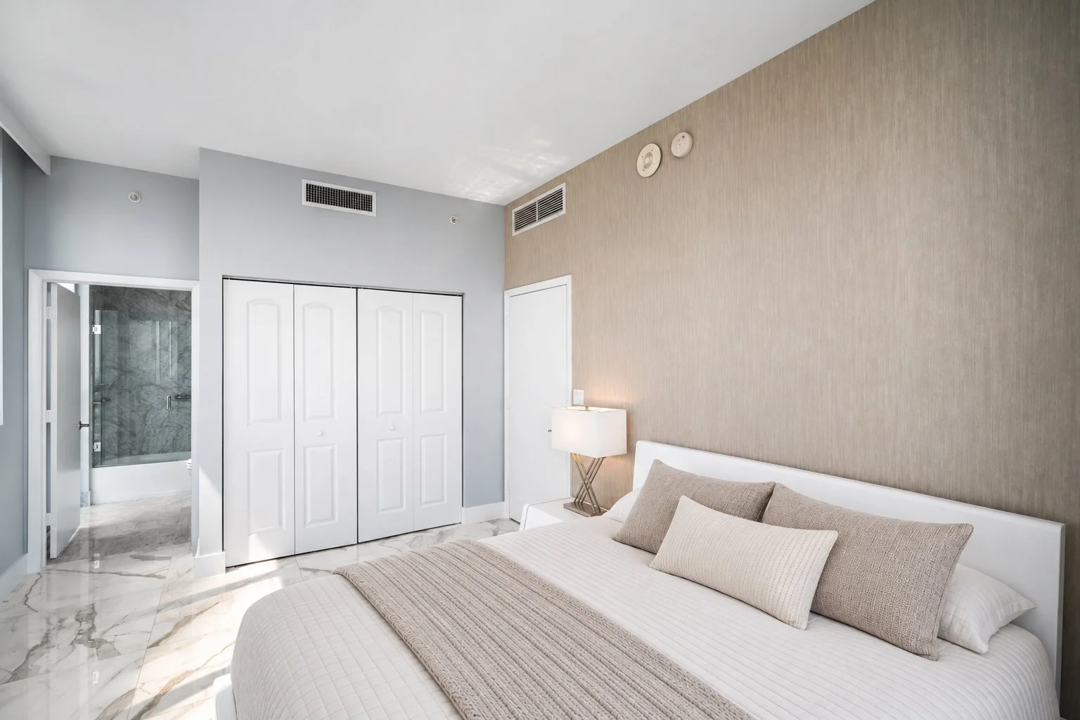 Modern bedroom with a large bed, beige and white throw pillows, a beige textured accent wall, a white bedside table with a white lamp, a closed white closet door, and an open door to a bathroom with marble walls.