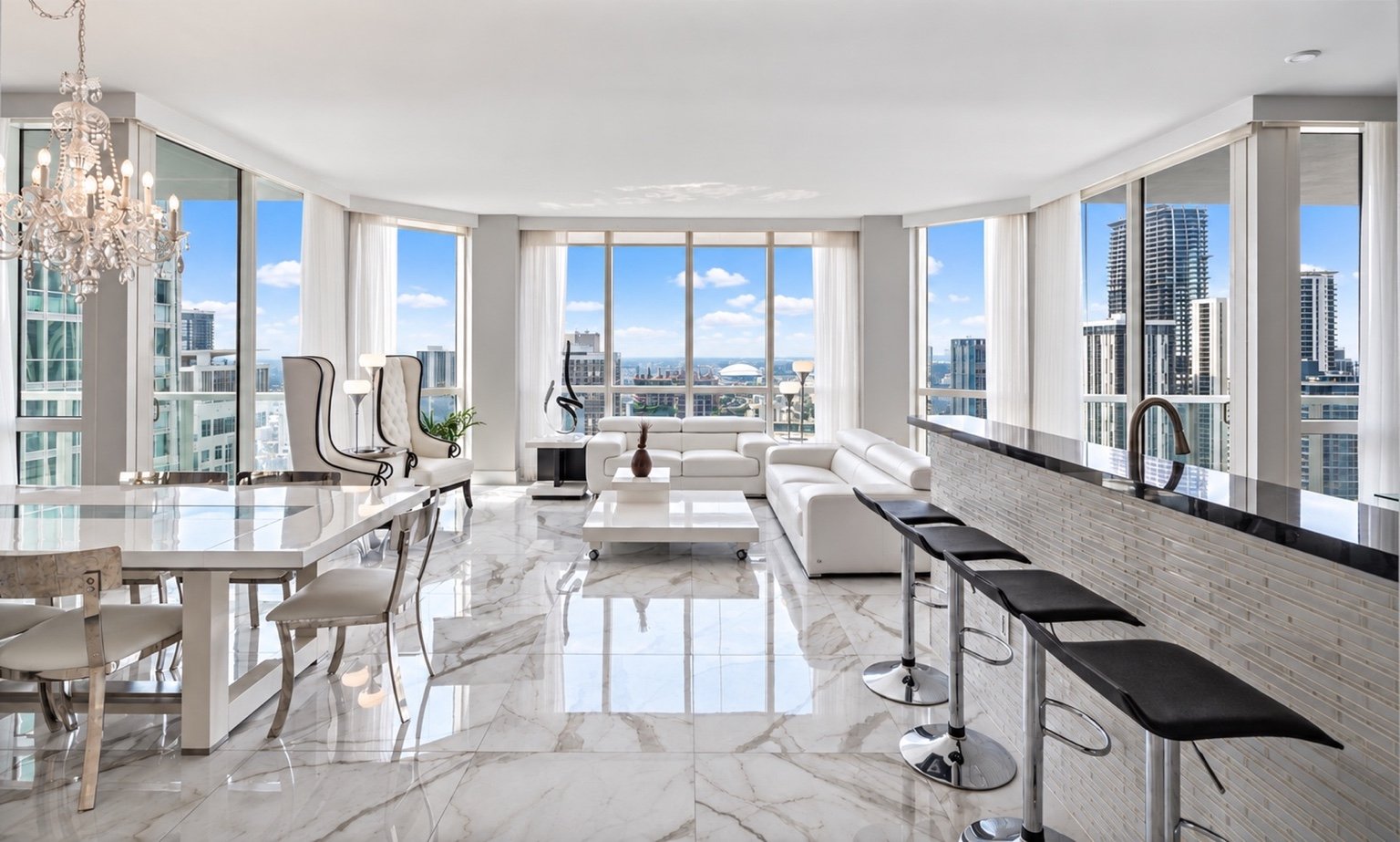 Modern high-rise apartment living room with white furniture, marble floors, large windows with city views, dining area, and kitchen bar with black stools.